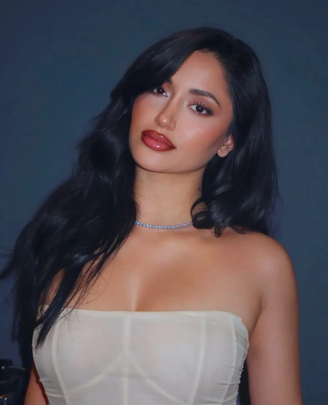 Woman with long black hair, wearing strapless beige top and a silver necklace, against dark background.