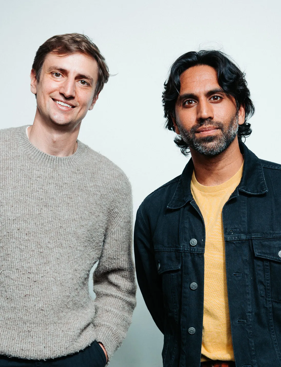 Two men standing side by side, one smiling in a gray sweater, the other with dark hair in a black jacket.