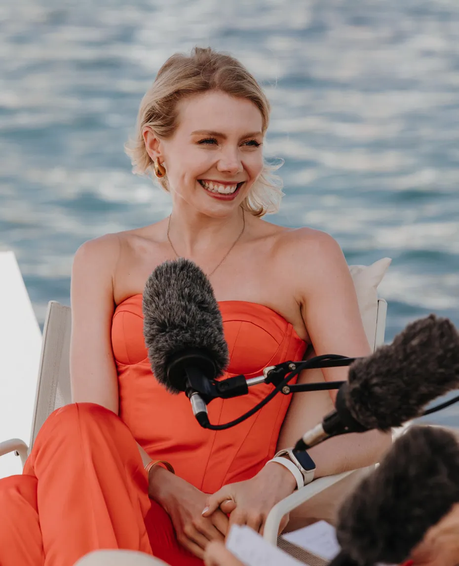 Smiling woman in an orange dress sitting and speaking into microphones by the water.