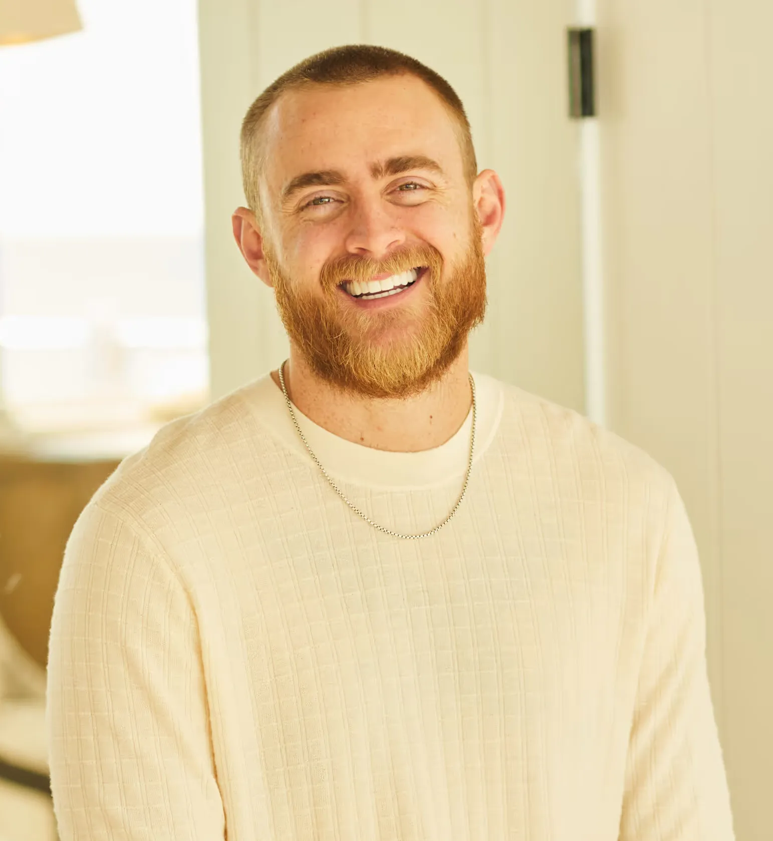 Smiling man with red beard in a cream sweater and silver chain standing indoors.