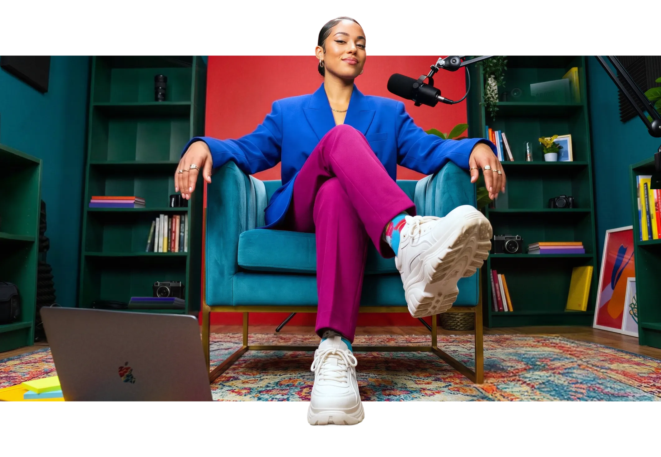 Confident woman in blue blazer and purple pants sits cross-legged in teal chair with mic and laptop nearby.