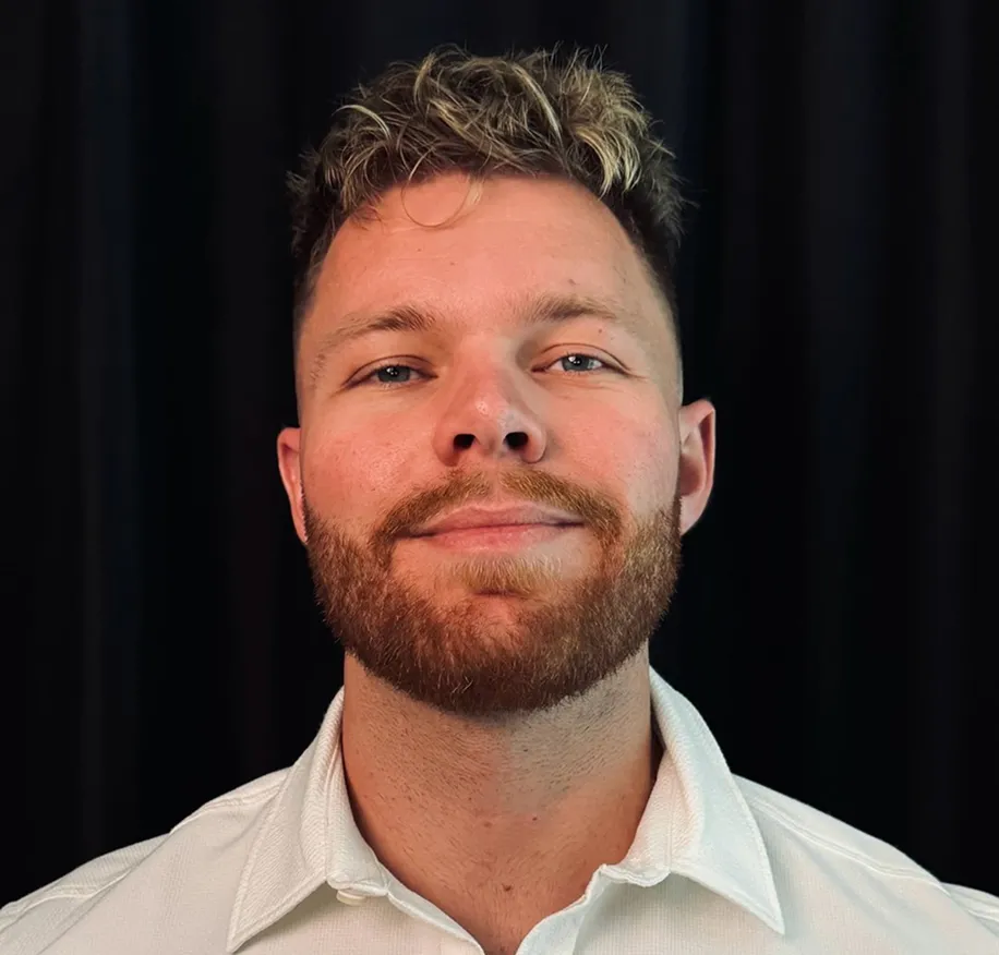 Smiling man with trimmed beard and curly blond hair wearing a white collared shirt against black background.