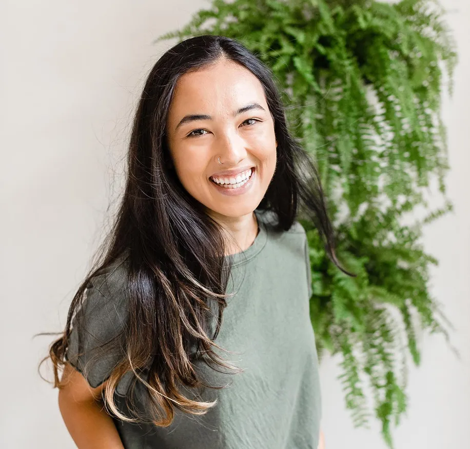 Smiling woman with long dark hair and nose ring wearing green shirt with green plant background.