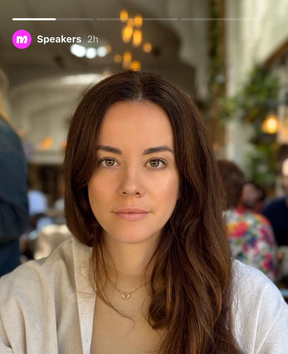 Woman with long brown hair wearing a beige top and necklace seated in a softly lit restaurant.