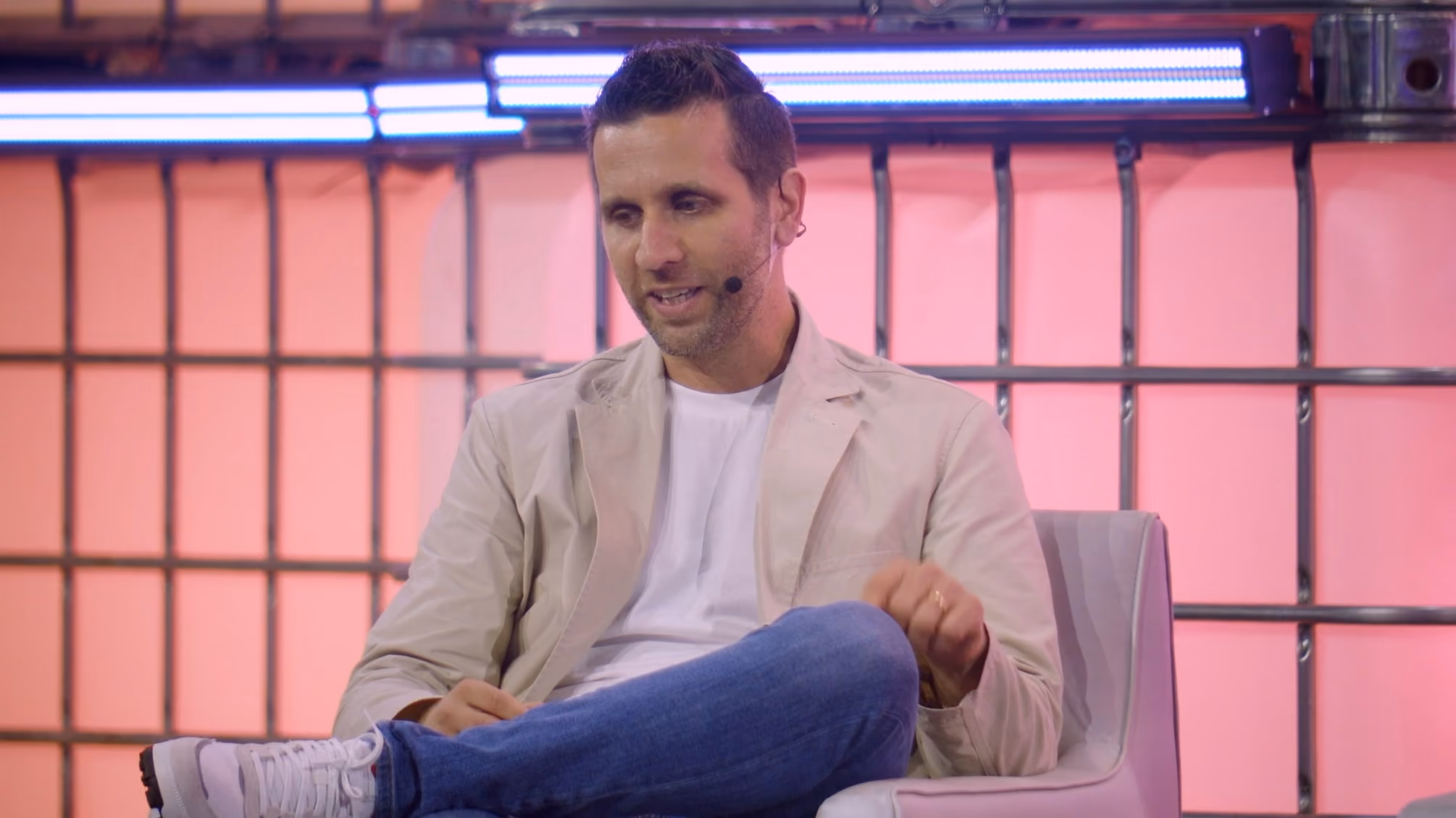 Man wearing a beige jacket and blue jeans seated on a chair with a microphone on his cheek.