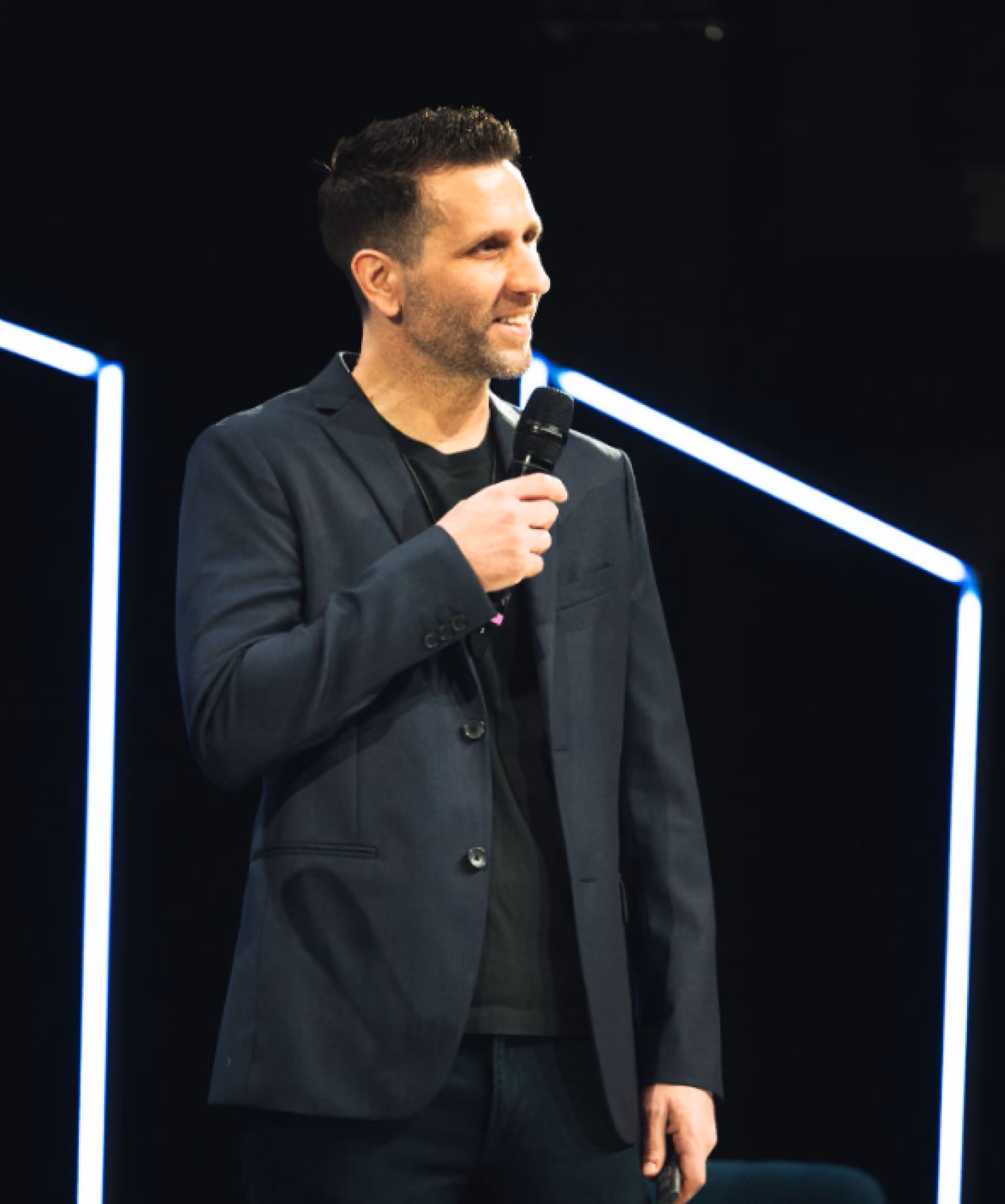 Man in dark blazer holding a microphone speaking on stage with lit geometric lines behind him.