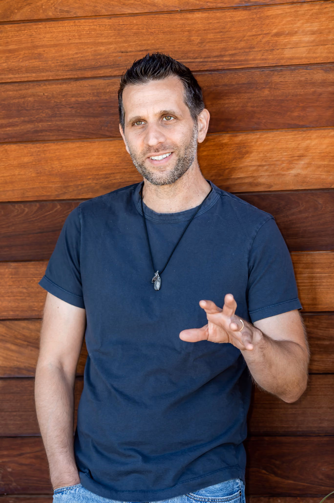 Man with short dark hair and stubble wearing a black shirt and necklace gestures with his right hand.