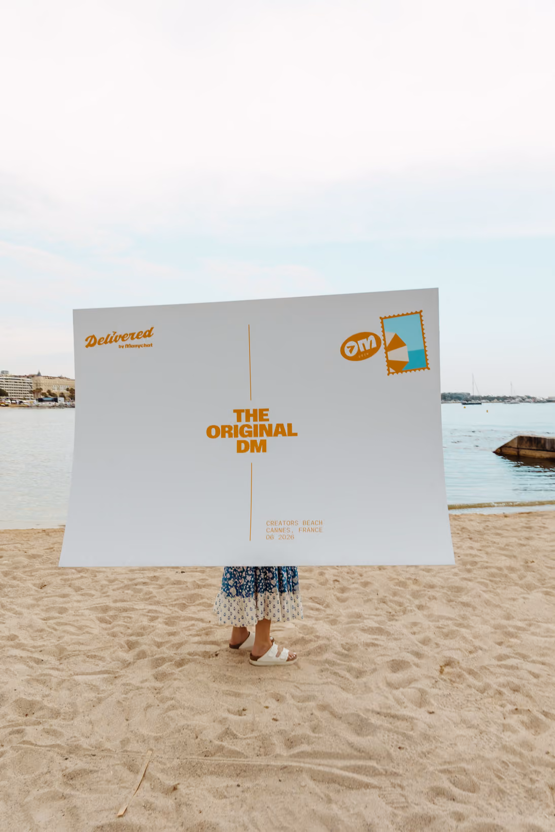 Person on sandy beach holding large postcard sign reading 'The Original DM' with water in background.