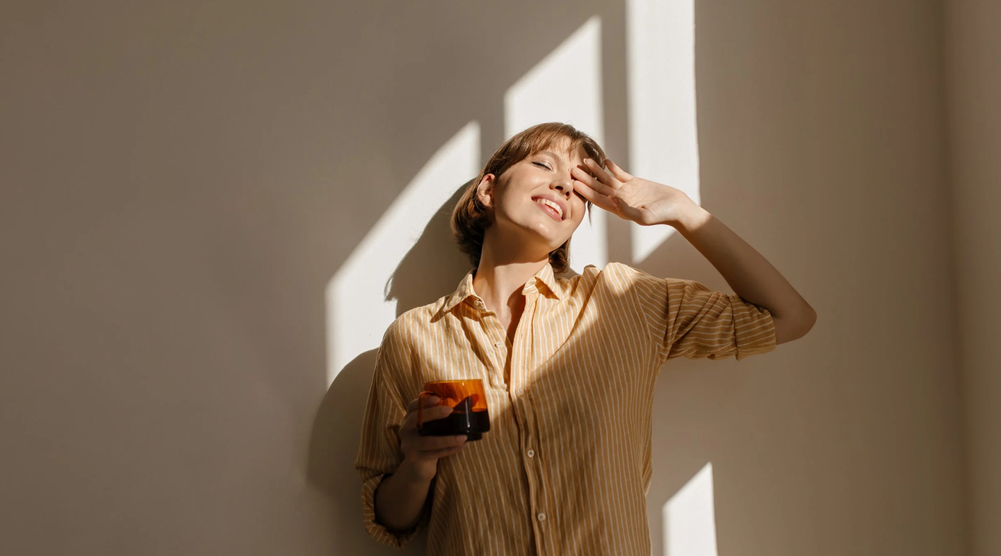 Woman in striped beige shirt holding a cup and smiling with eyes closed while shielding her face from sunlight.