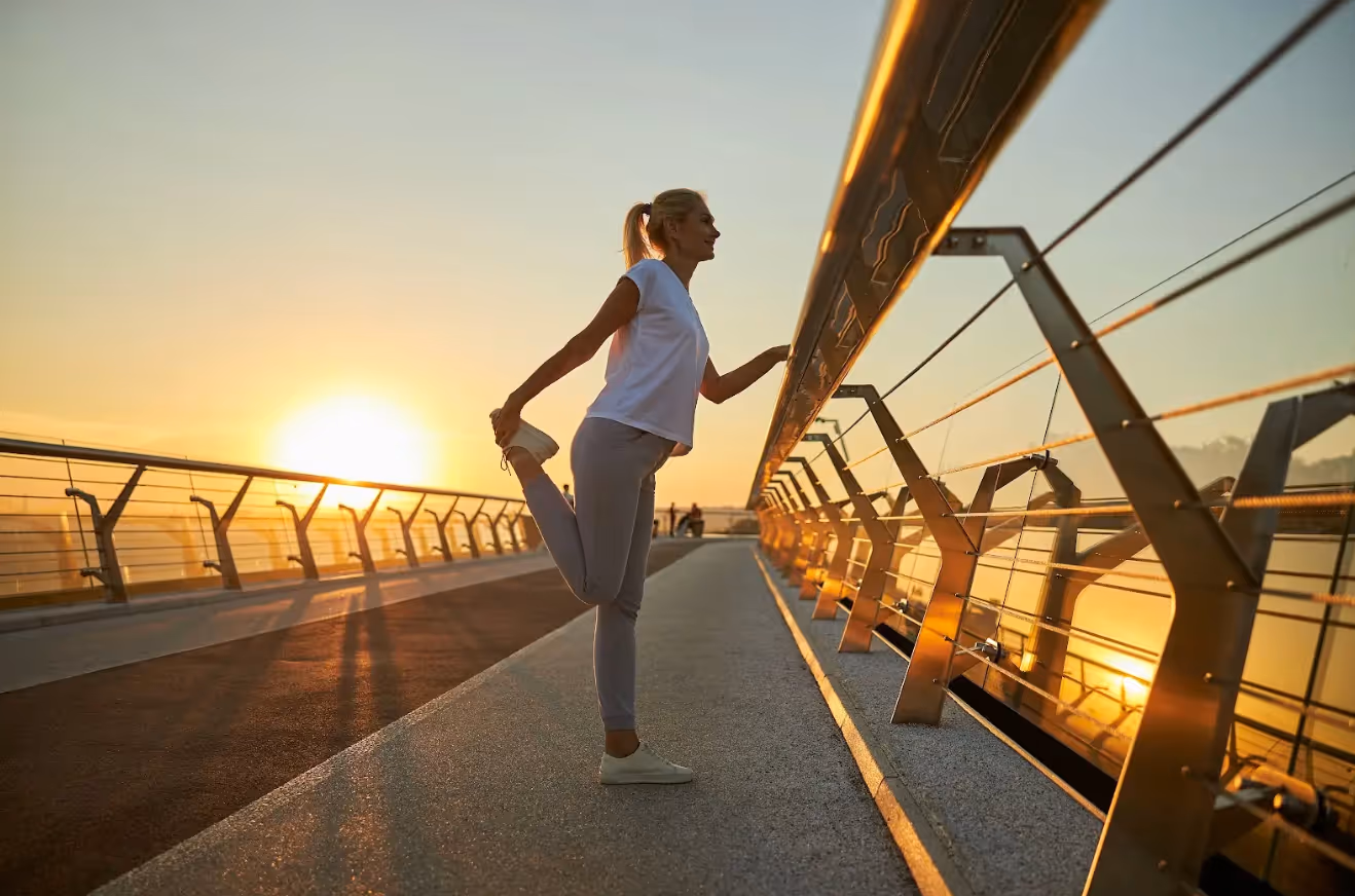 Woman in athletic wear stretching her leg on a bridge at sunrise.