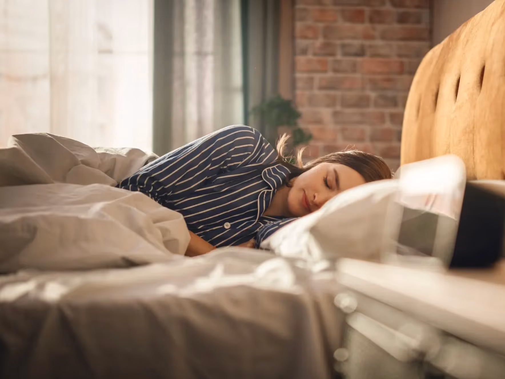 Young woman peacefully sleeping on her side in bed with striped pajamas and beige bedding.