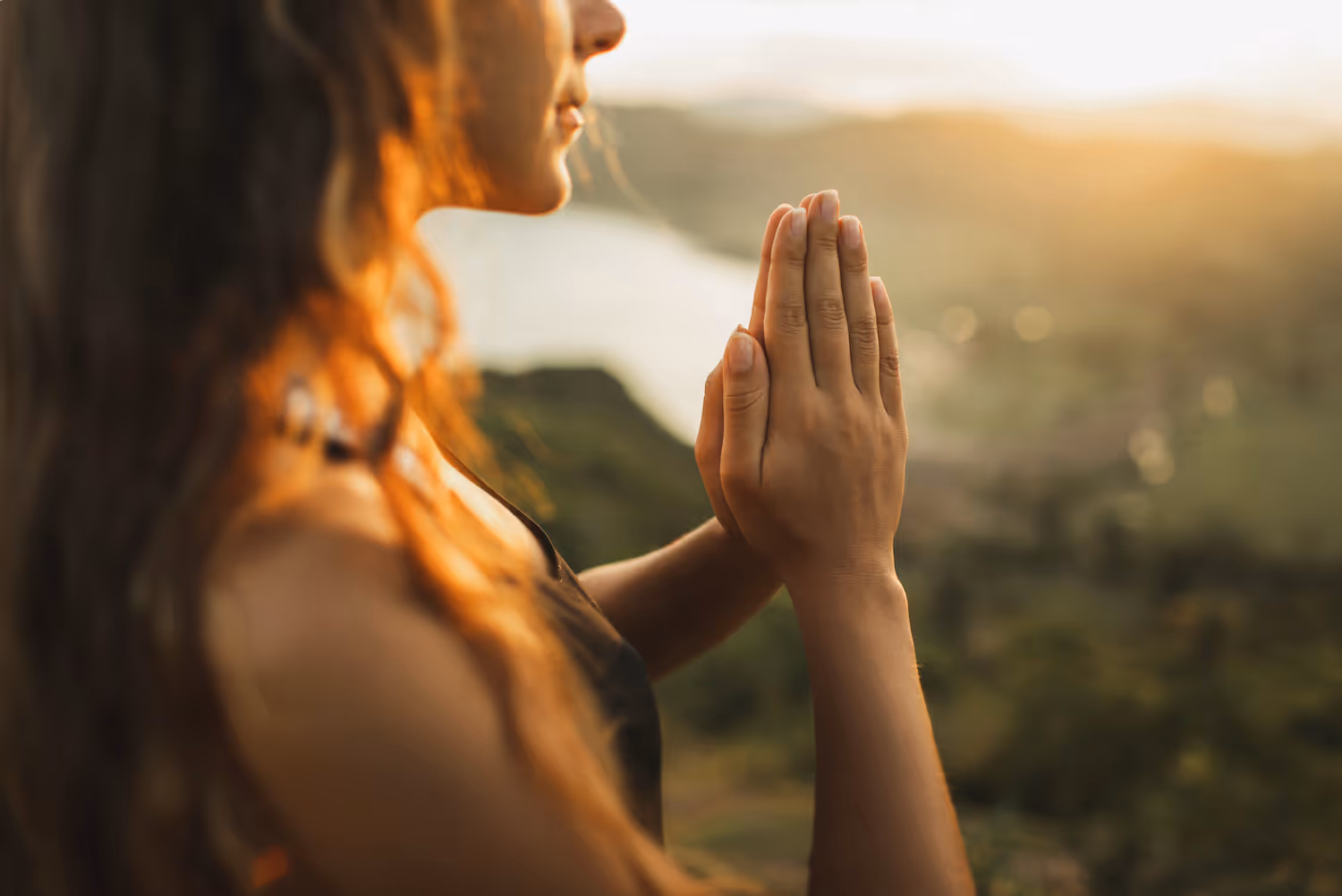 Woman with long hair holding hands in prayer or meditation with a blurred natural landscape and sunlight in the background.