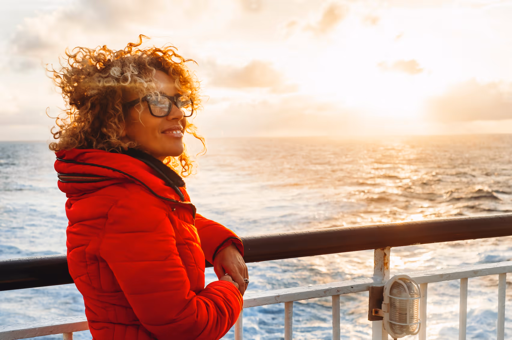 Woman in red jacket with curly hair and glasses leaning on a ship railing, watching the ocean at sunset.