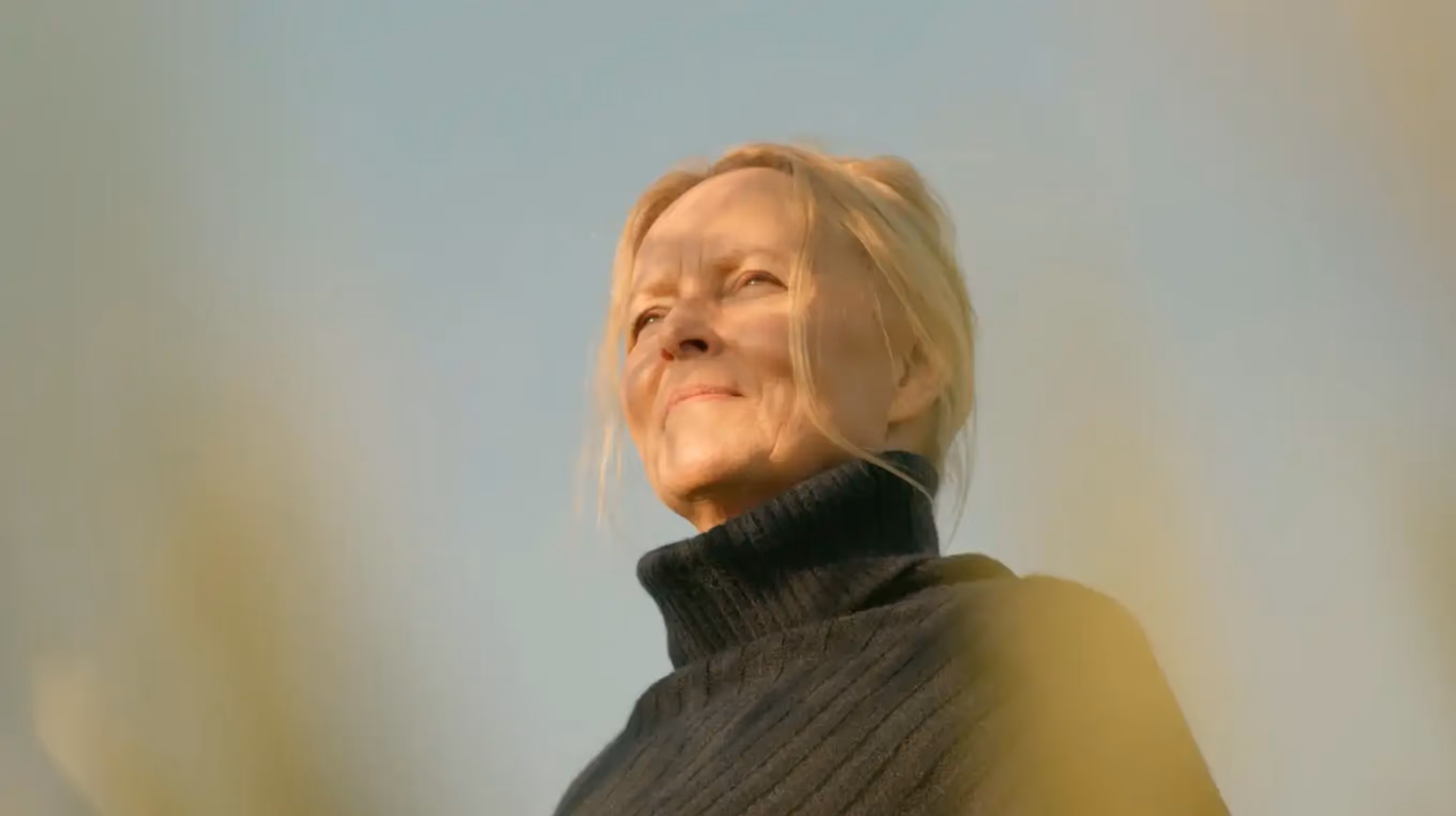 Woman with blonde hair wearing a black turtleneck sweater, looking thoughtfully into the distance against a clear sky.