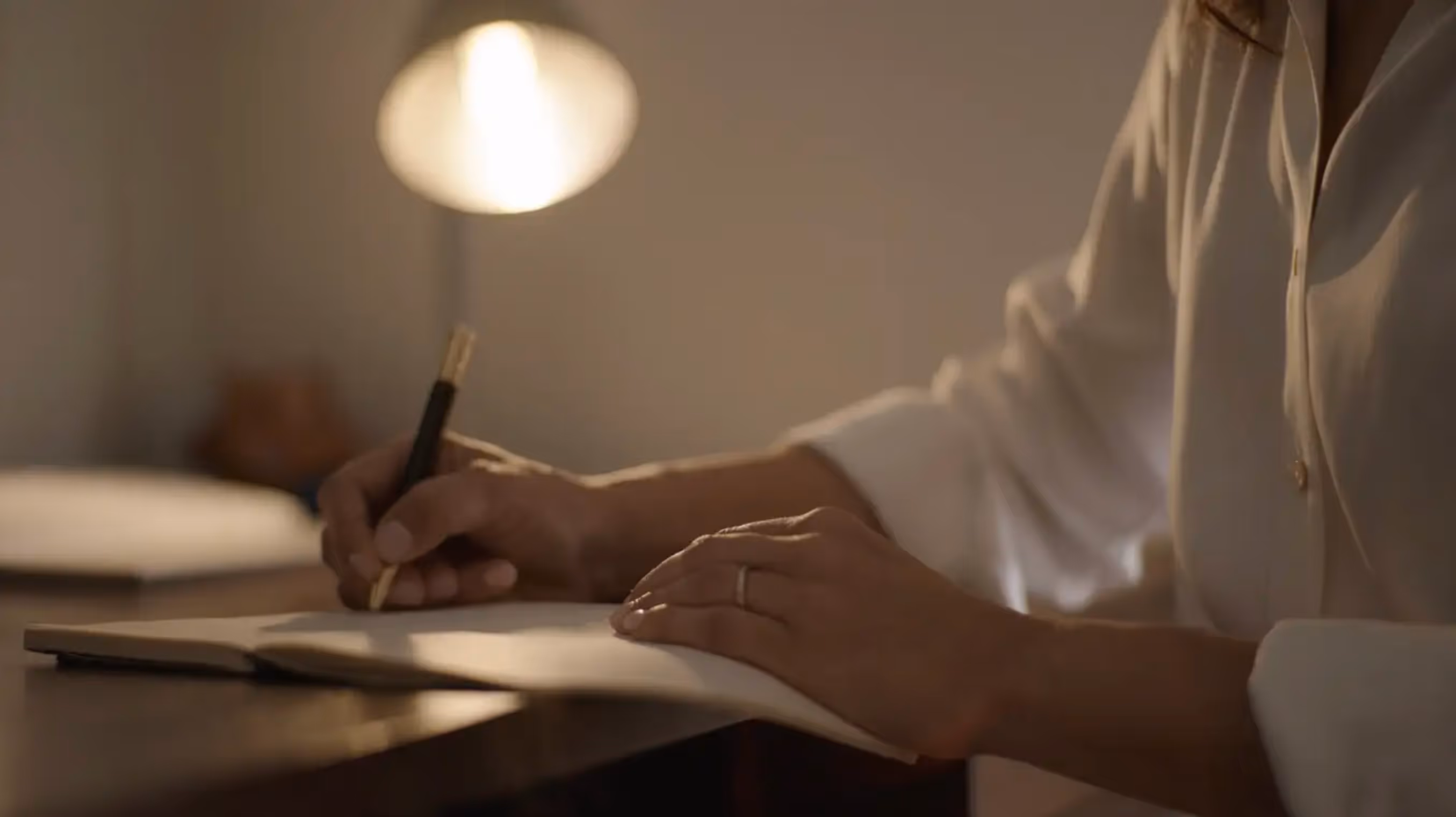 Person wearing a white shirt writing in a notebook at a wooden desk with a lamp in the background.