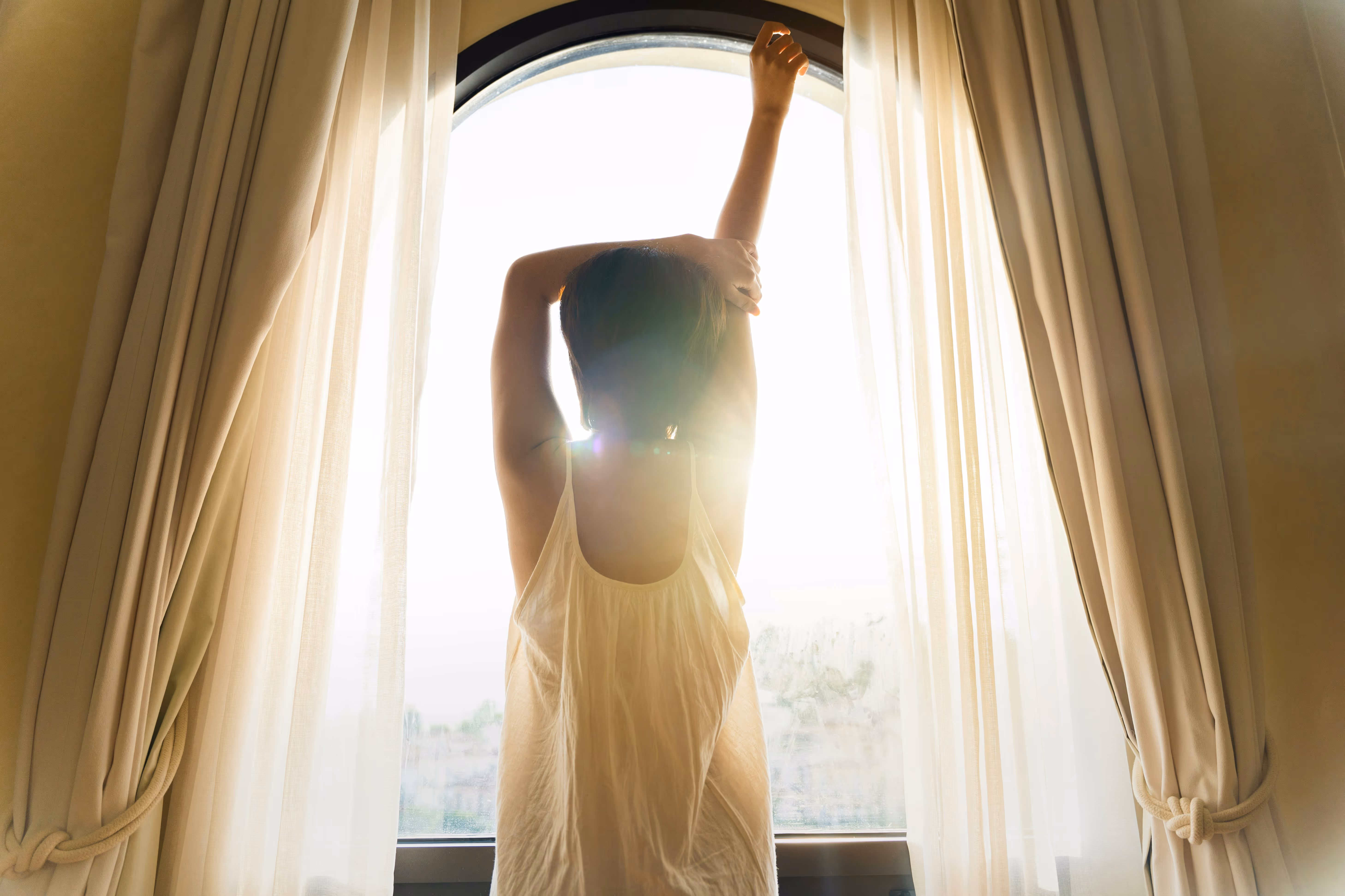 Woman stretching her arms while standing in front of a large arched window with sunlight shining through sheer curtains.
