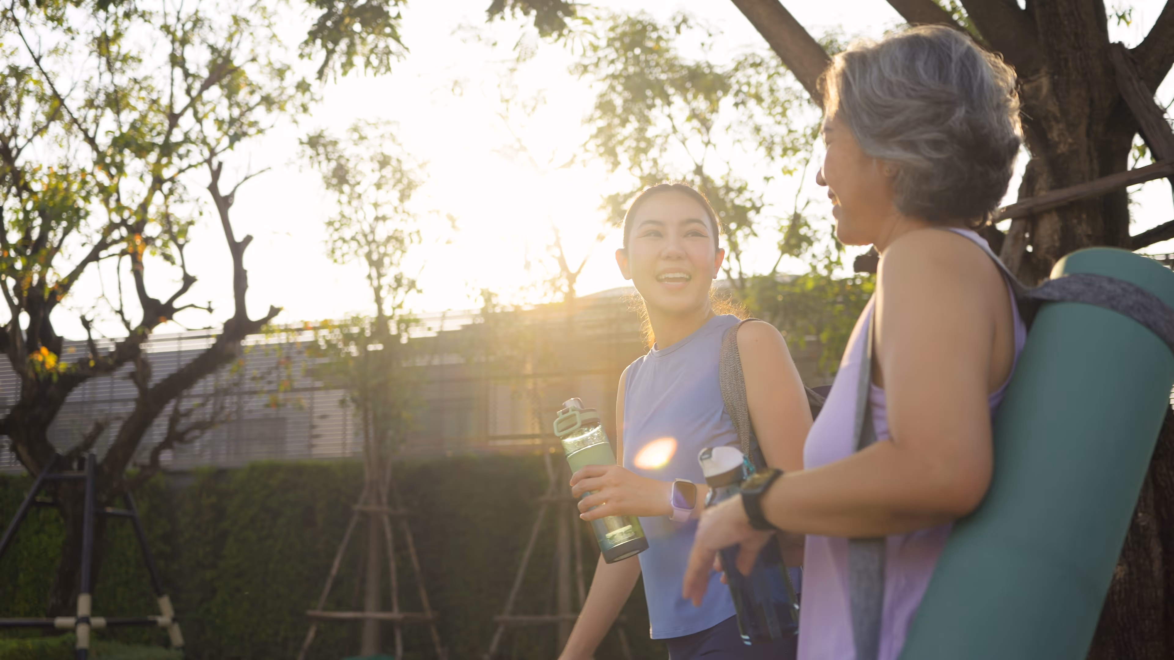 Two women in athletic wear holding water bottles and smiling outdoors with trees and sunlight behind them.