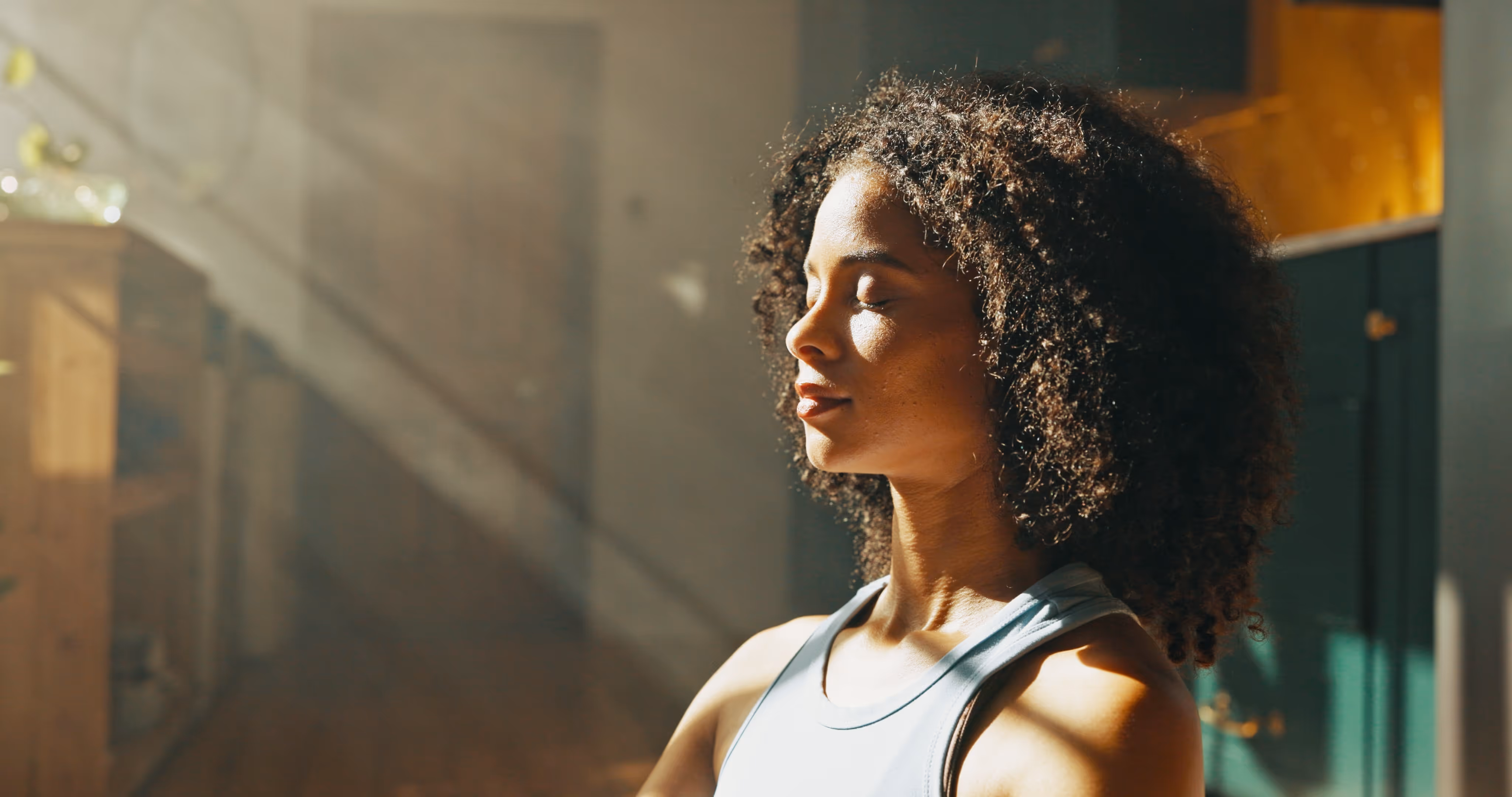 Woman with curly hair meditating indoors with eyes closed and sunlight streaming in.
