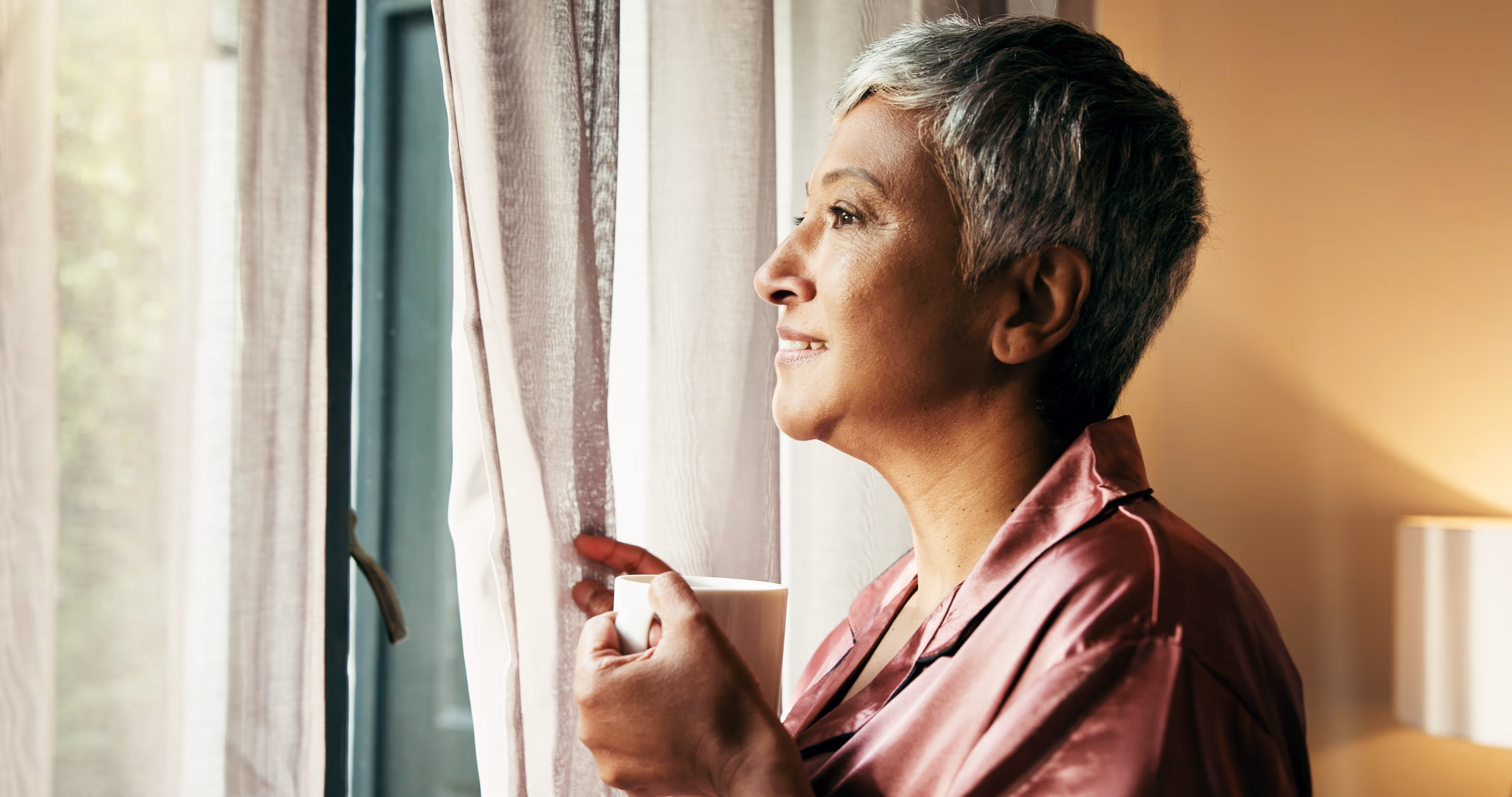 Smiling mature woman in pink satin pajamas holding a white mug while looking out a window with sheer curtains.