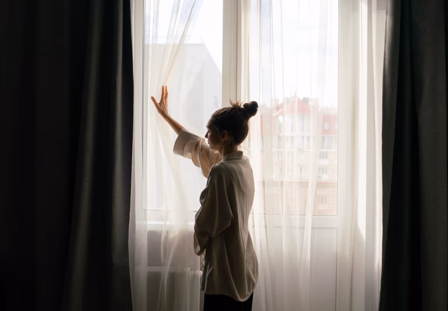 Woman in a light robe standing by a window, gently touching the sheer curtain with city buildings visible outside.
