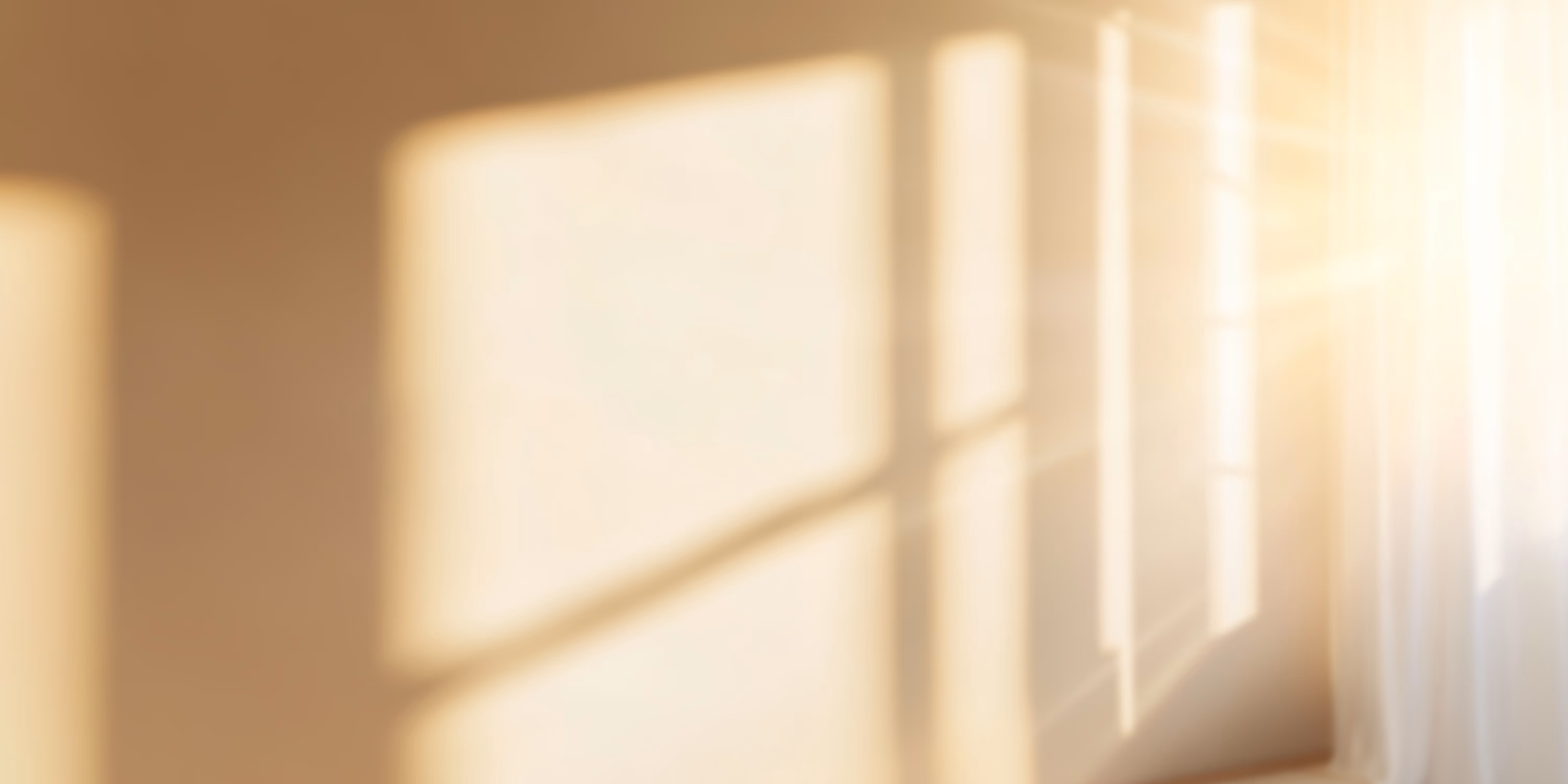 Soft sunlight streaming through a window casting warm, blurred shadows on a beige wall with sheer white curtains on the side.