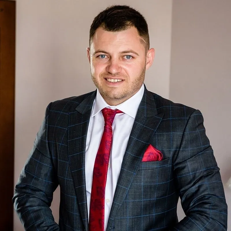 Man in a dark checkered suit, white shirt, and red tie smiling at the camera against a neutral background.