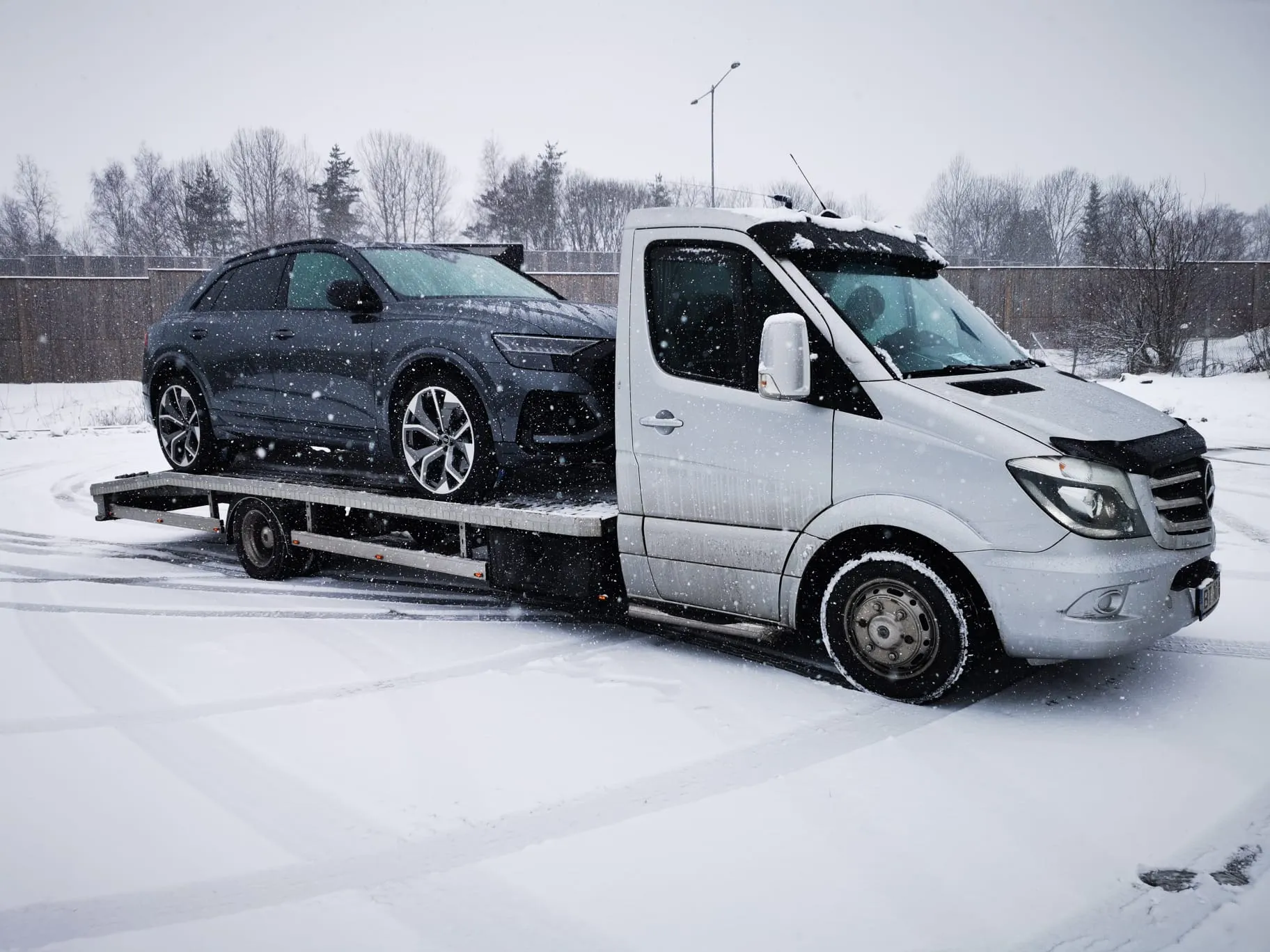 White flatbed tow truck carrying a black SUV in a snowy parking lot with trees and a wooden fence in the background.