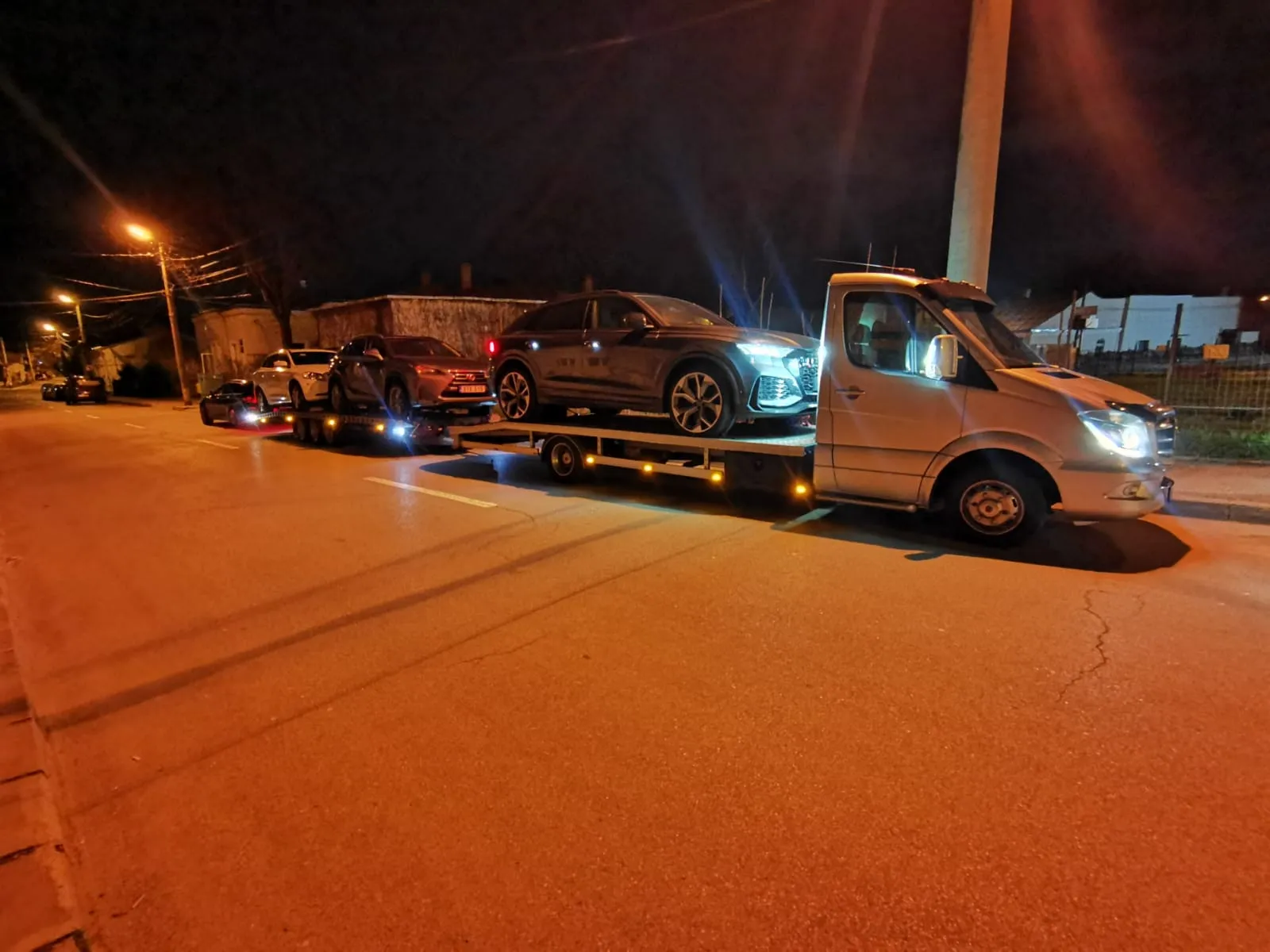 Three cars being transported on tow trucks parked on a street at night under streetlights.