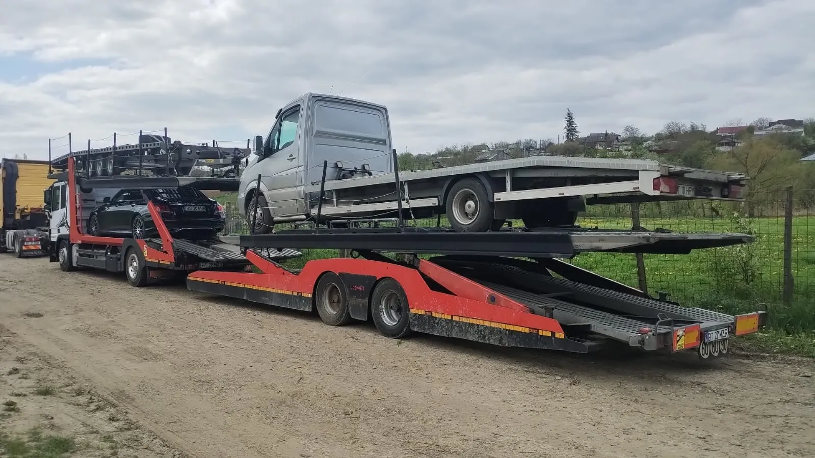 Car transporter truck loaded with multiple vehicles, including a black car and a silver flatbed truck, parked on a dirt road with grassy hills and houses in the background.