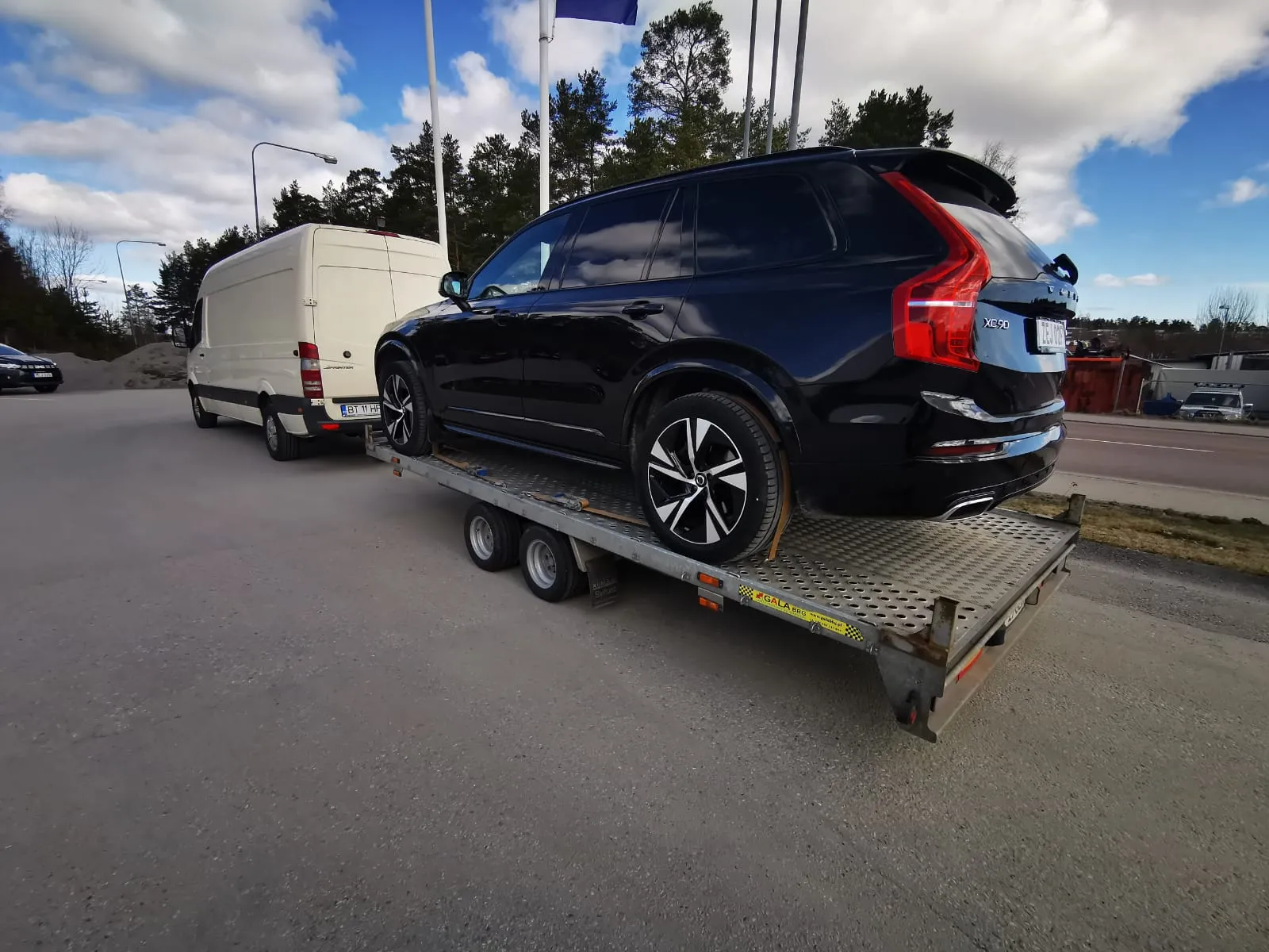 Black Volvo XC90 SUV loaded on a flatbed trailer attached to a white van on a road beside trees and buildings.