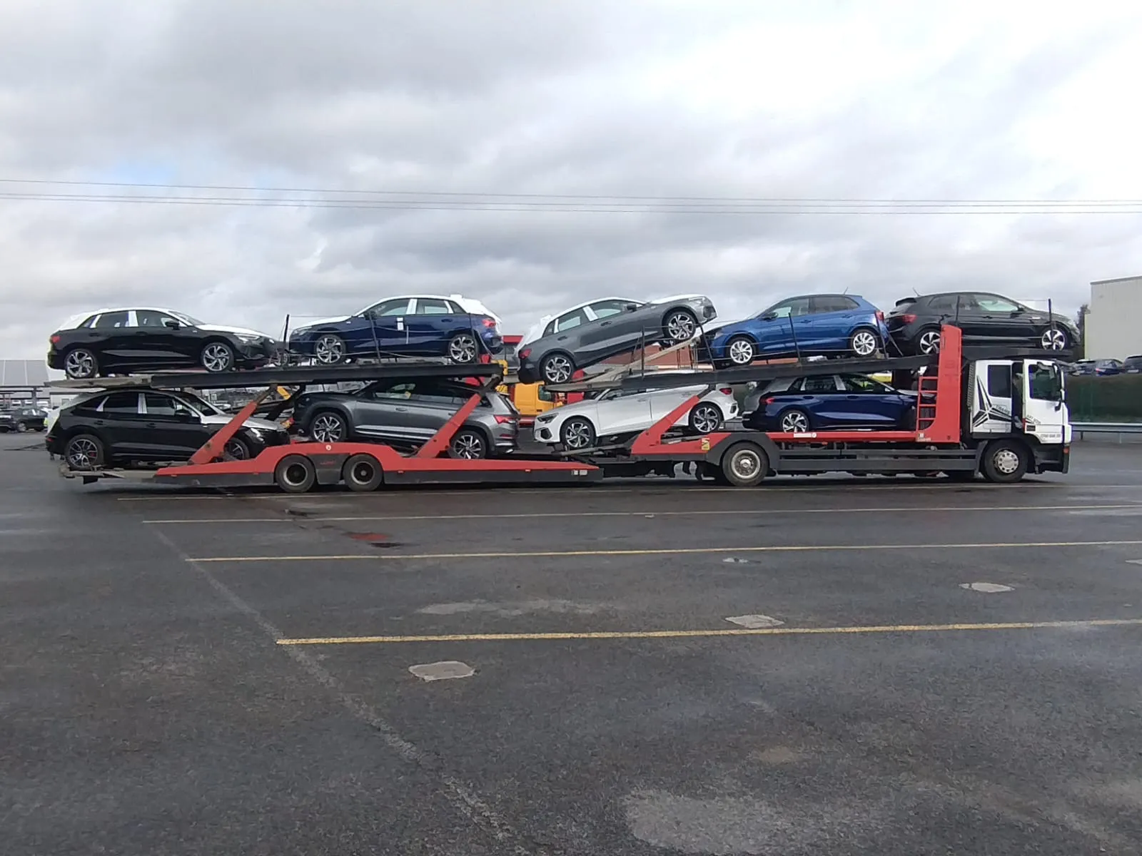 Car carrier truck parked in a lot, loaded with eight new cars in various colors including black, blue, gray, and white.