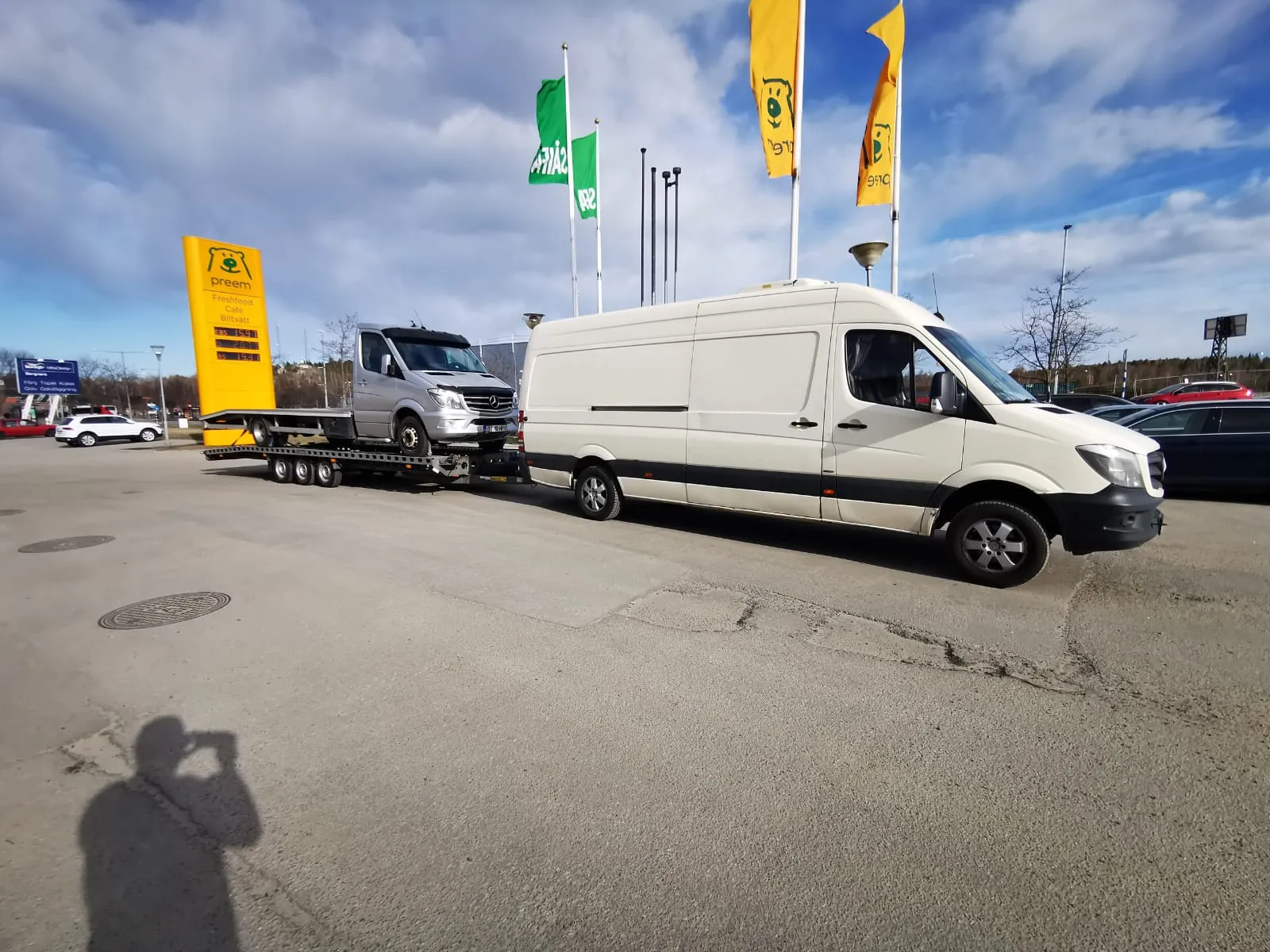 White van towing a flatbed trailer carrying a silver Mercedes van in a parking lot under a blue sky.