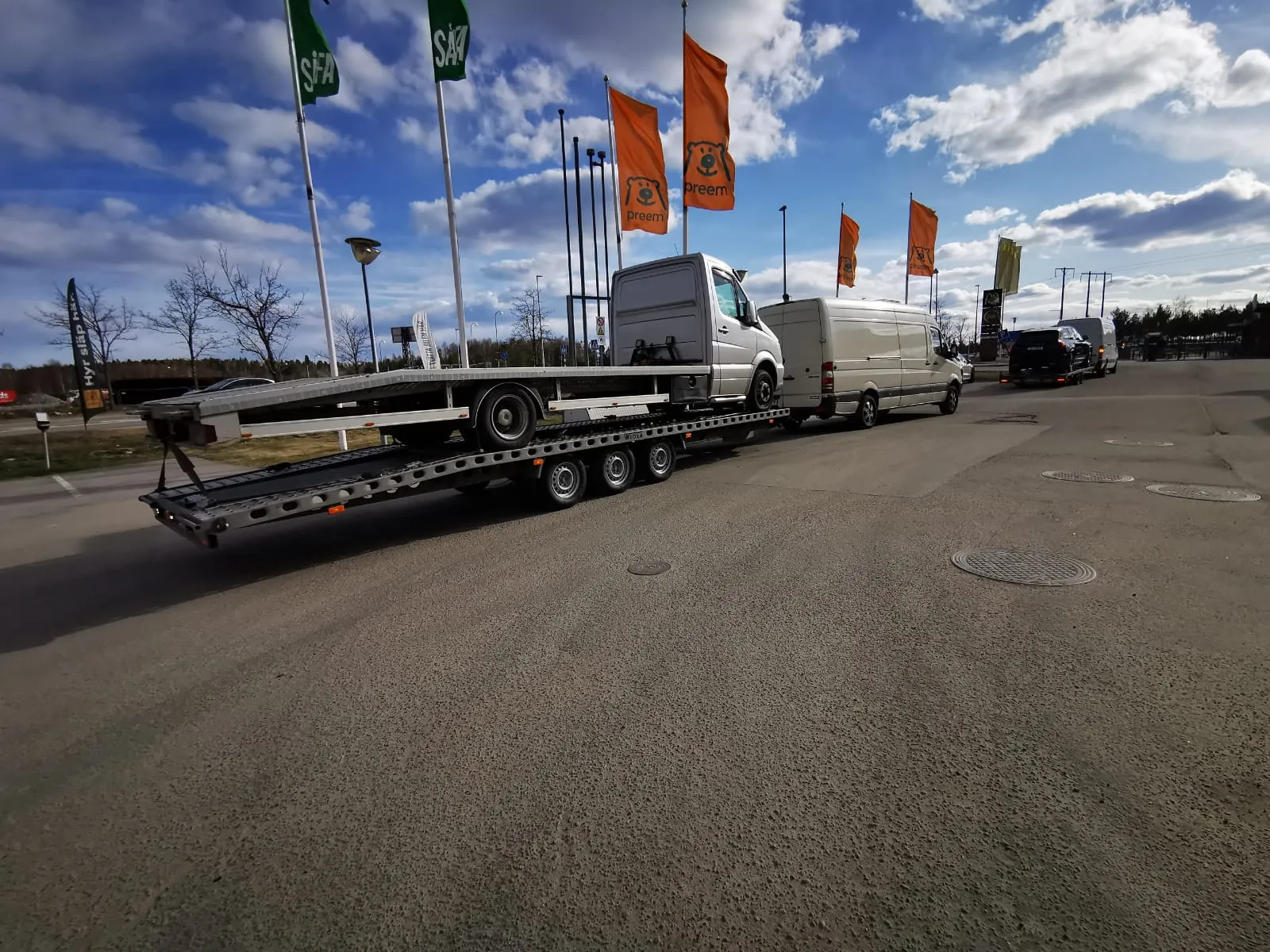 White cargo vans parked in a row on a large trailer in an open lot under a partly cloudy blue sky.