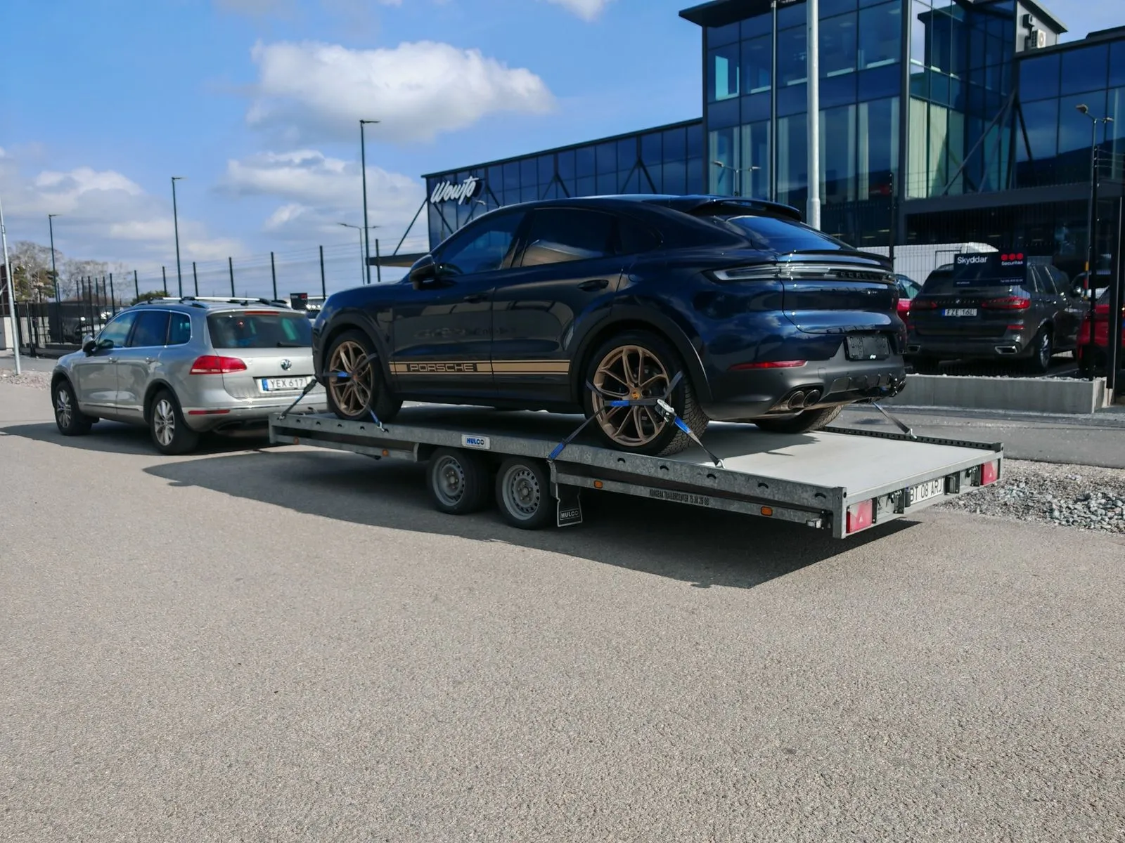 Blue Porsche SUV secured with straps on a flatbed trailer hitched to a silver SUV on a street near modern buildings.
