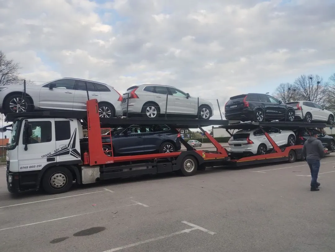 Car carrier truck loaded with six SUVs parked in an outdoor lot with a person walking nearby under a cloudy sky.