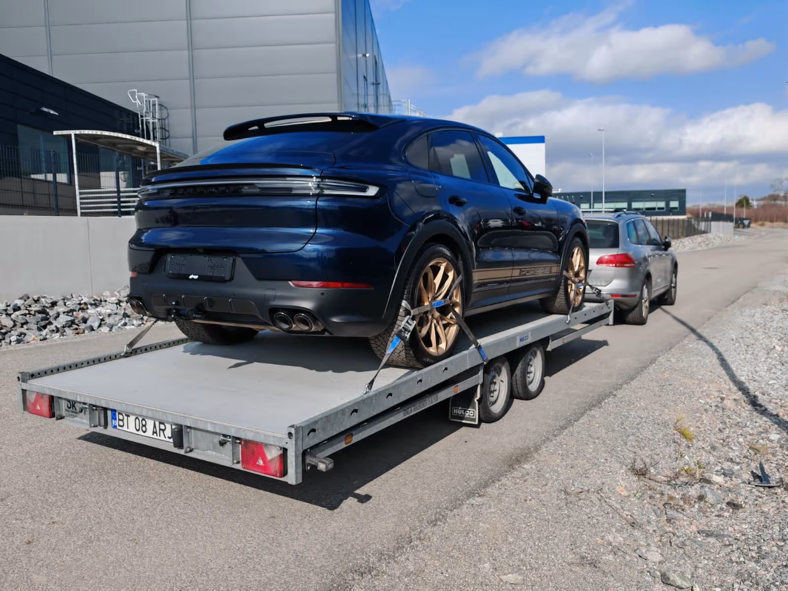 Dark blue Porsche SUV with gold rims secured on a flatbed trailer attached to a silver car on a paved road.
