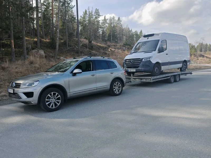 Silver SUV parked on a road in a forested area, towing a flatbed trailer carrying a white Mercedes van.