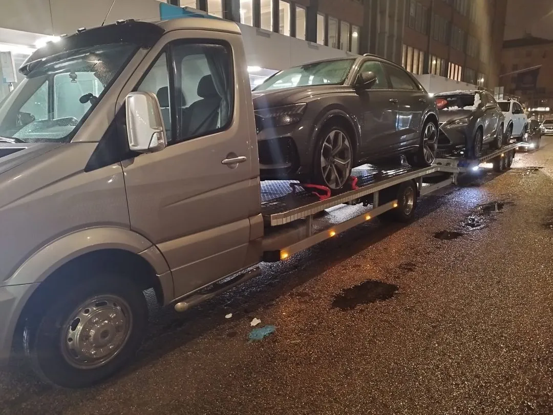 Flatbed tow truck carrying three cars parked on a wet city street at night.