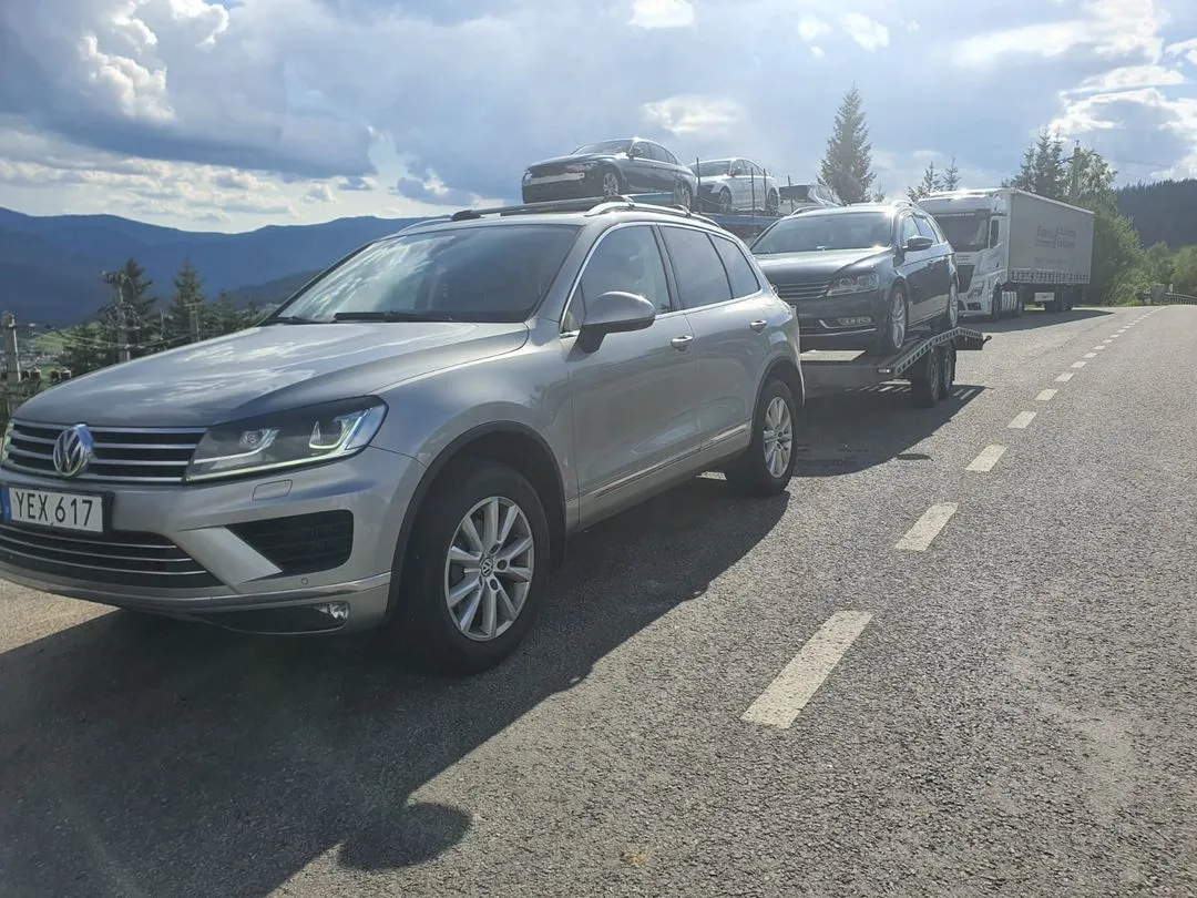 Silver Volkswagen SUV towing a car trailer loaded with two cars on a sunny mountain road.
