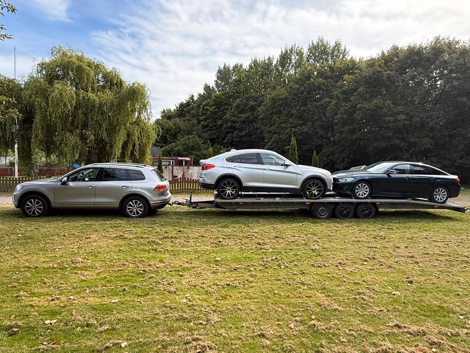 Silver SUV towing a trailer carrying a silver coupe and a black sedan on a grassy area with trees in the background.