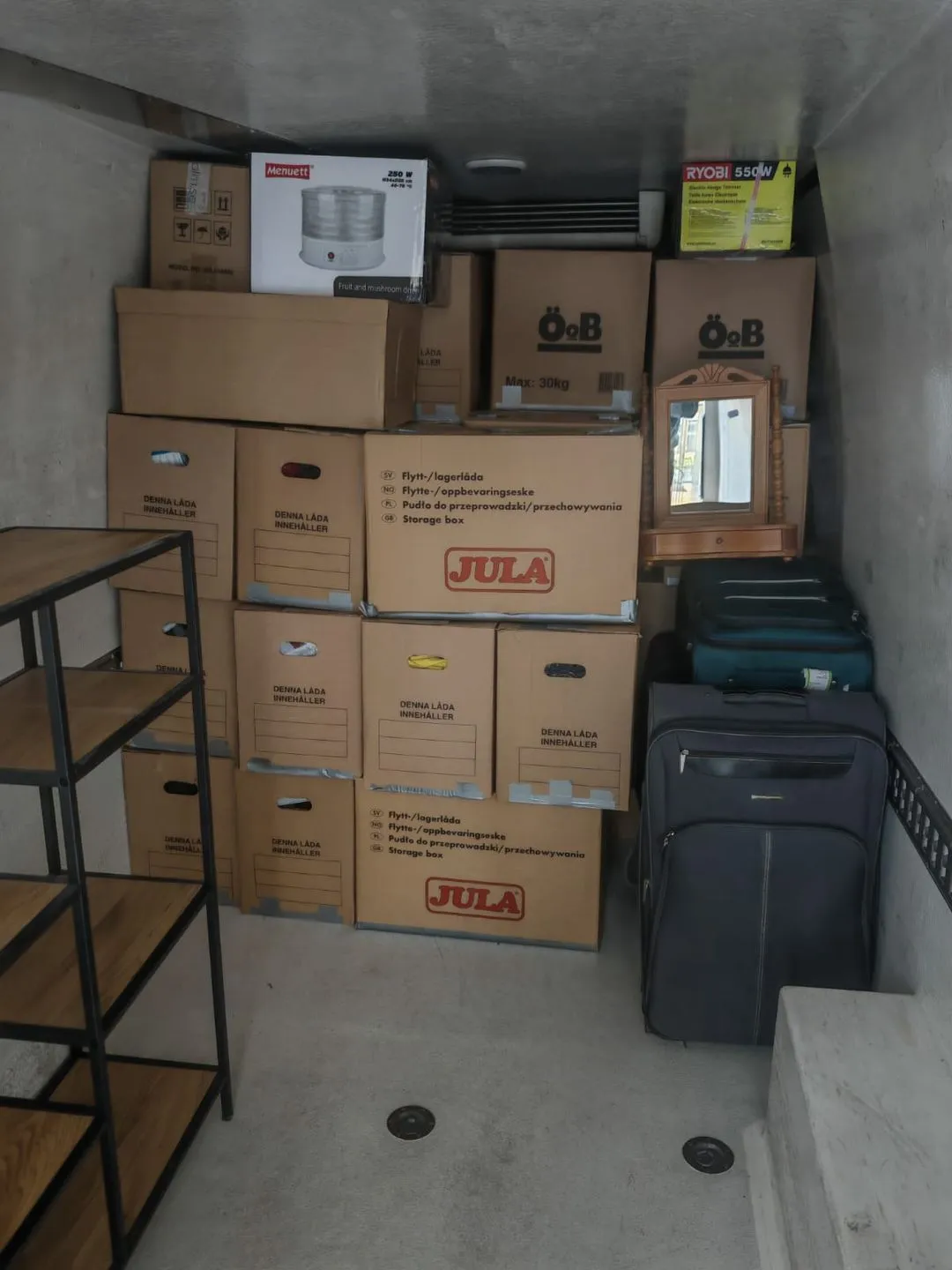 Stacked cardboard storage boxes, suitcases, a wooden shelf, and a small wooden mirror cabinet in a storage room.