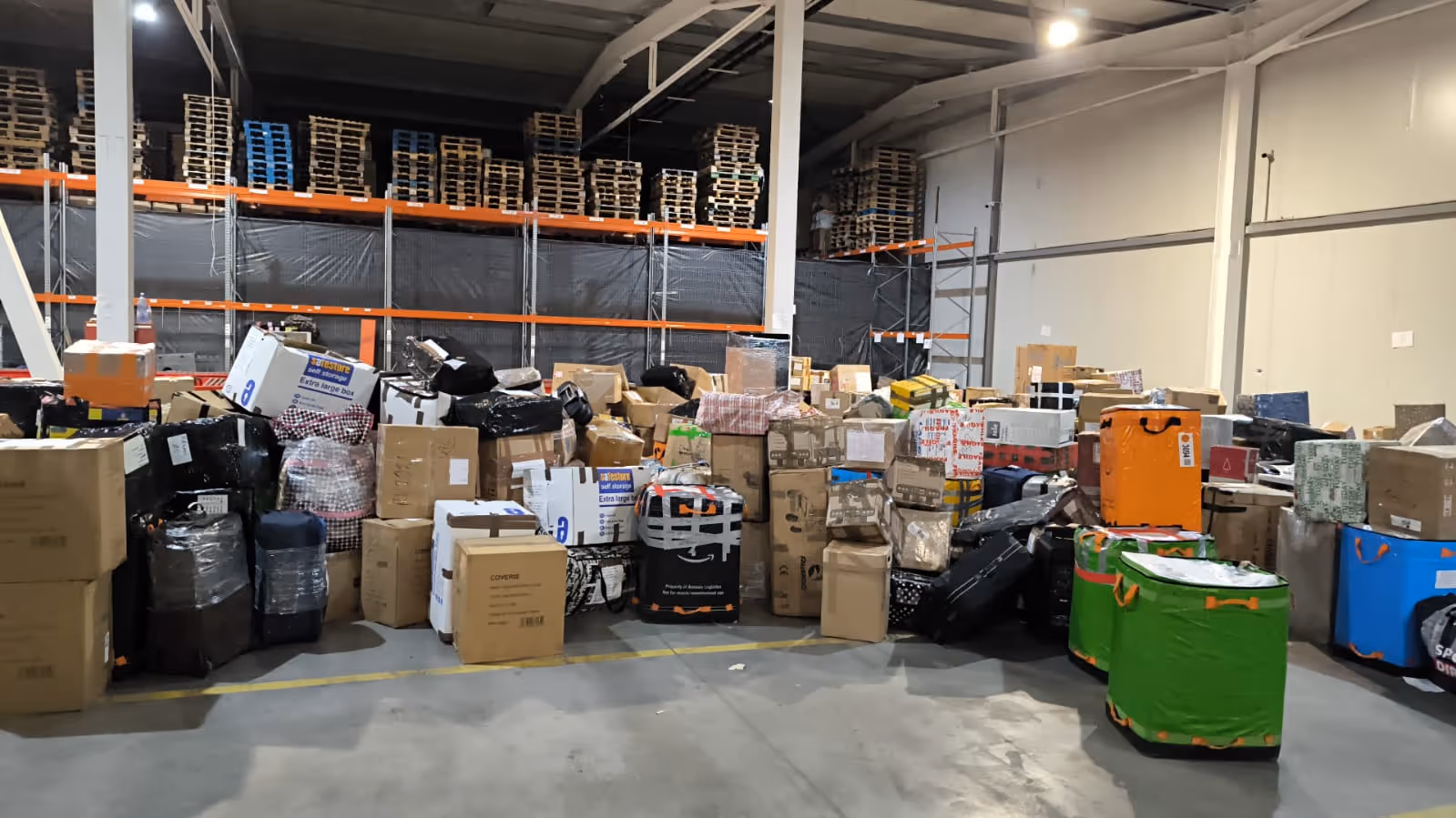 Warehouse interior with stacks of variously sized packed boxes and suitcases arranged on the floor in front of empty shelves and wooden pallets.