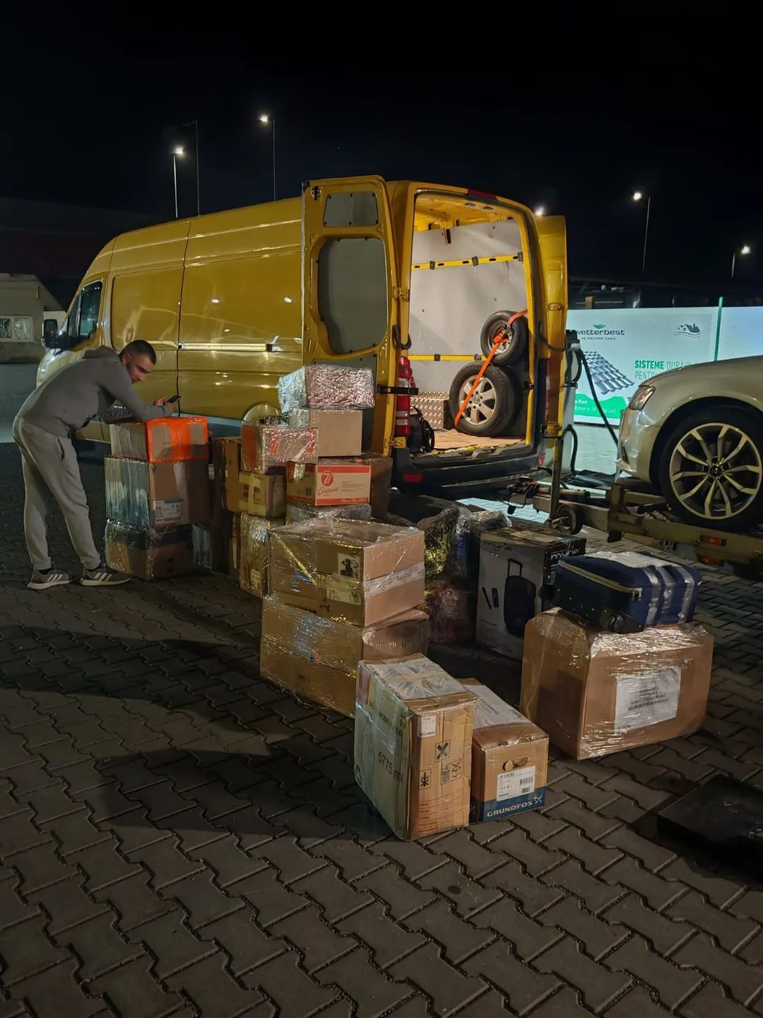 Man in gray tracksuit organizing packages outside an open yellow van at night.