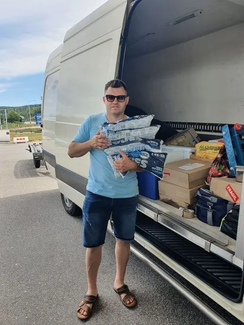 Man wearing sunglasses, blue t-shirt, and shorts holding several bags of ice in front of an open van loaded with boxes and bags.