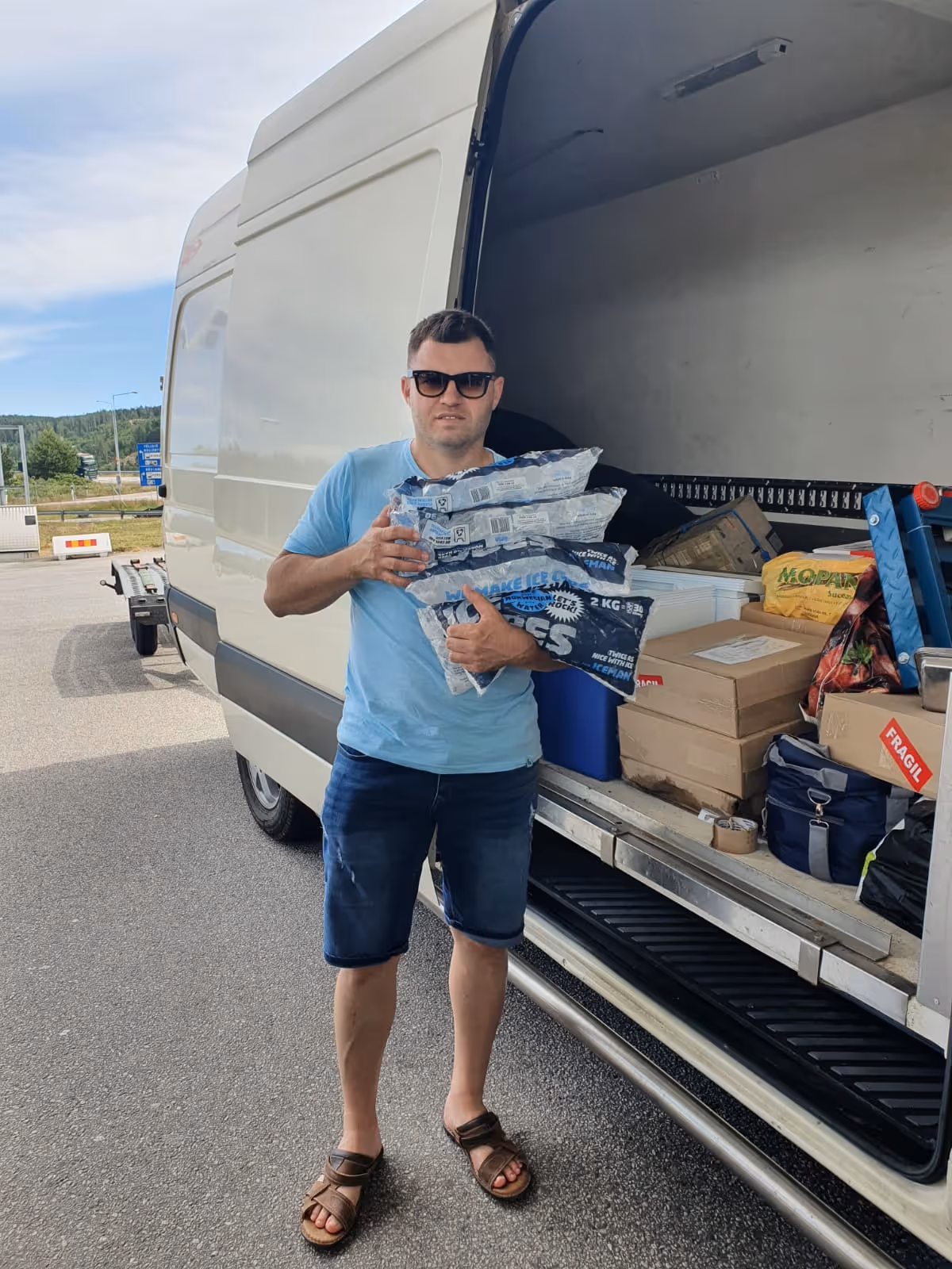 Man wearing sunglasses, blue t-shirt, and shorts standing next to an open white van holding several bags of ice.