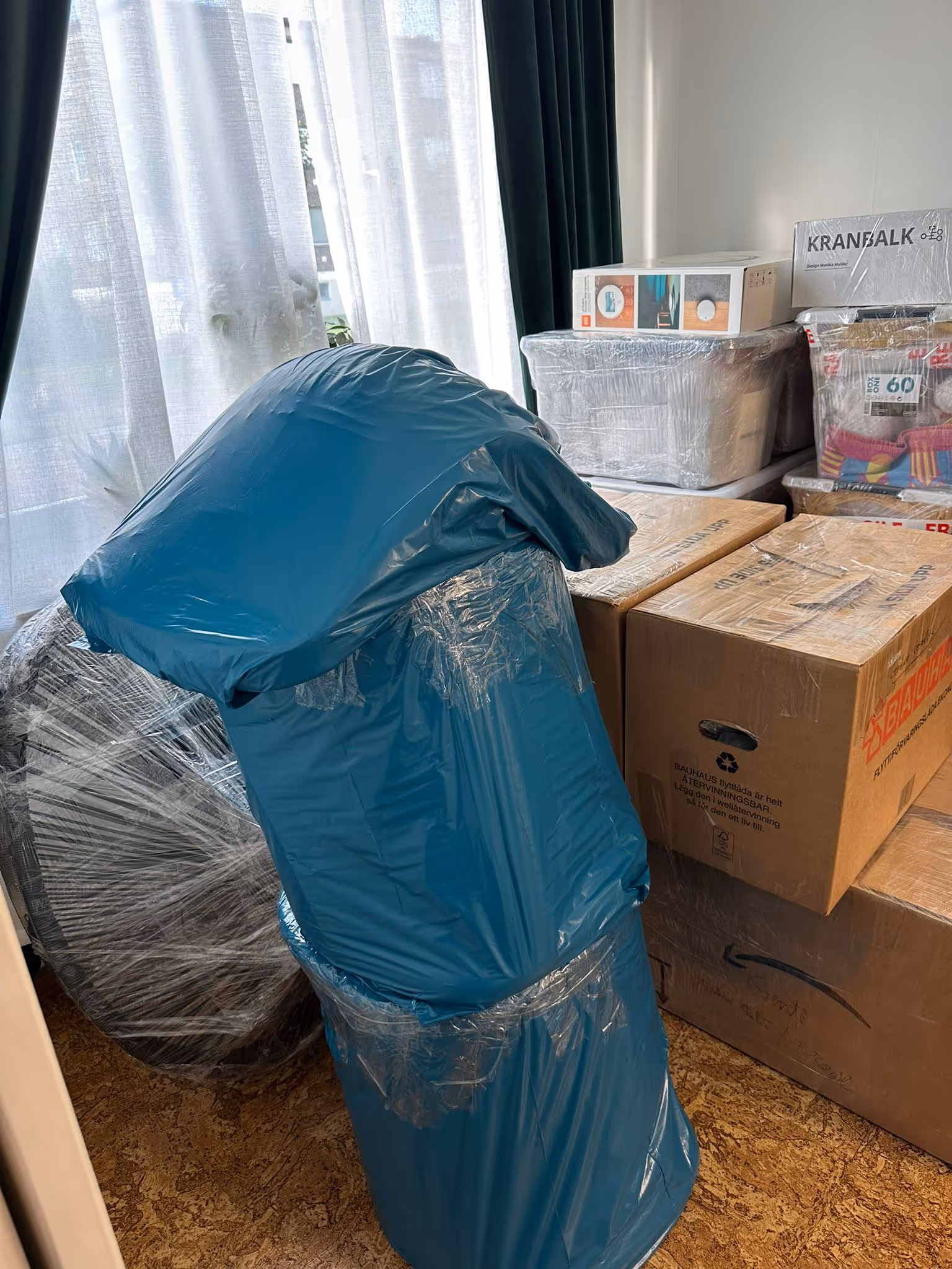Stack of packed moving boxes and wrapped furniture by a window with sheer curtains.
