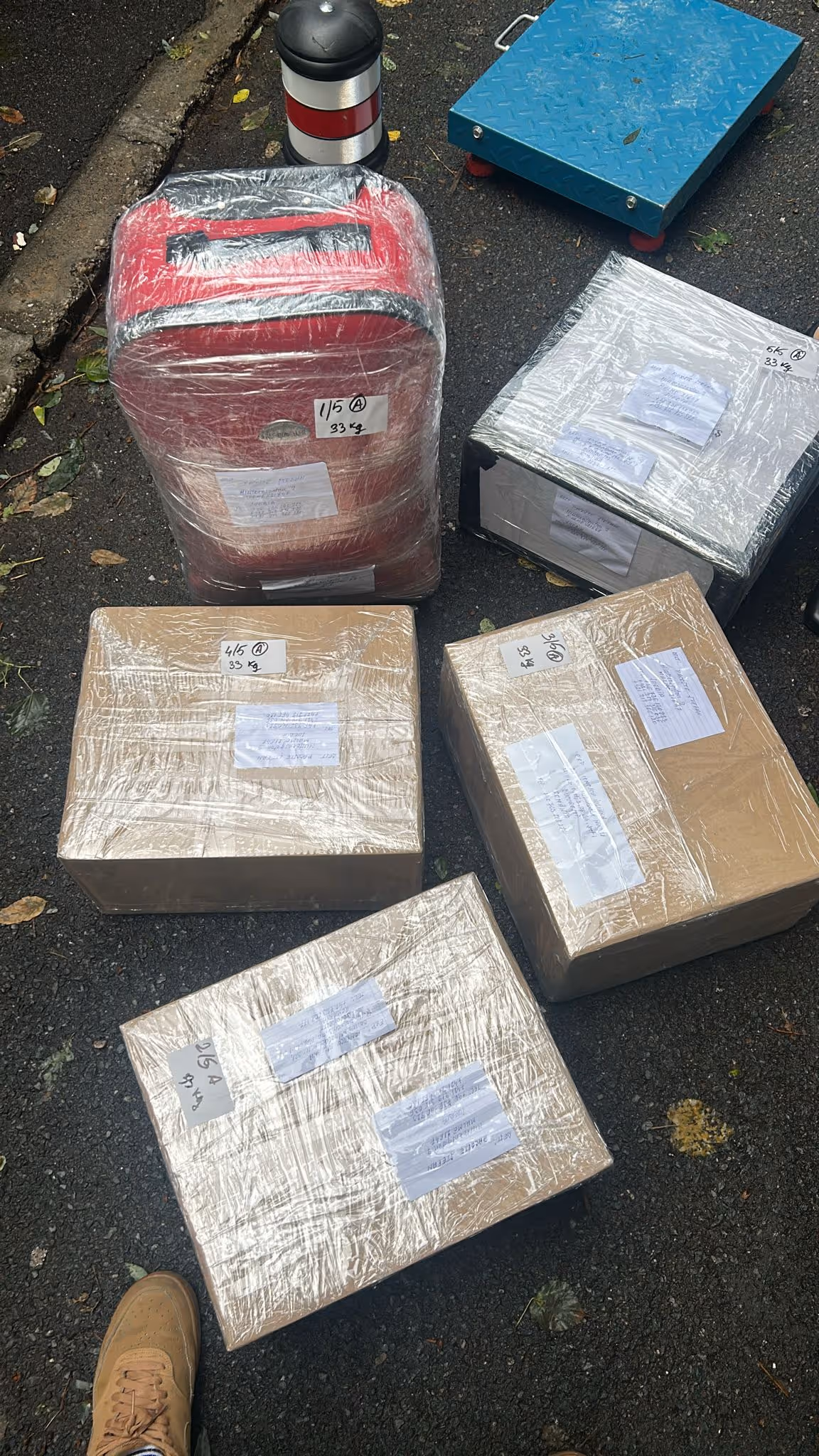 Five tightly wrapped packages with shipping labels placed on asphalt near a curb, including a red suitcase and four cardboard boxes.