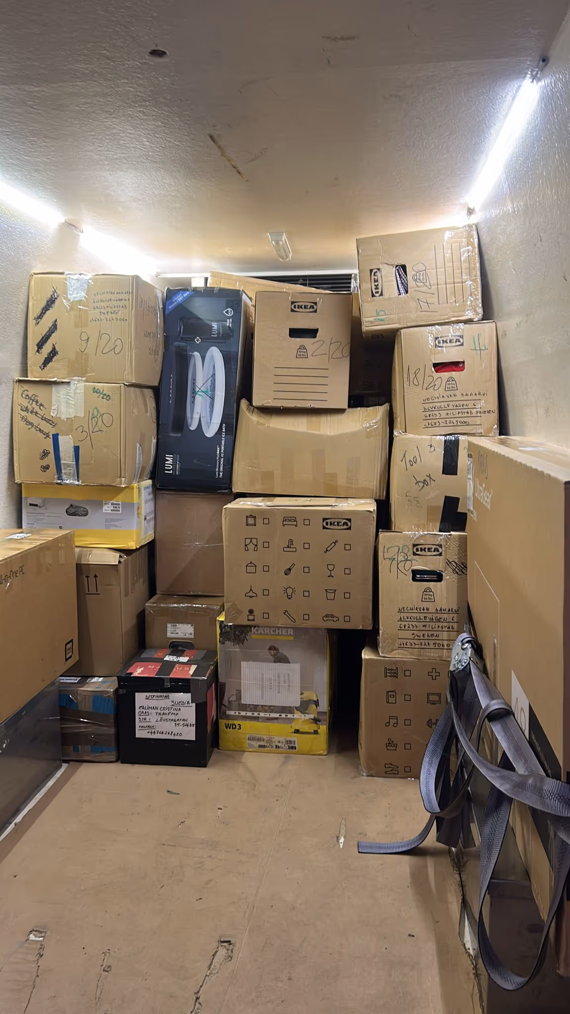 Stacked cardboard boxes and packages inside a delivery van, some labeled with IKEA logos and handwritten notes.