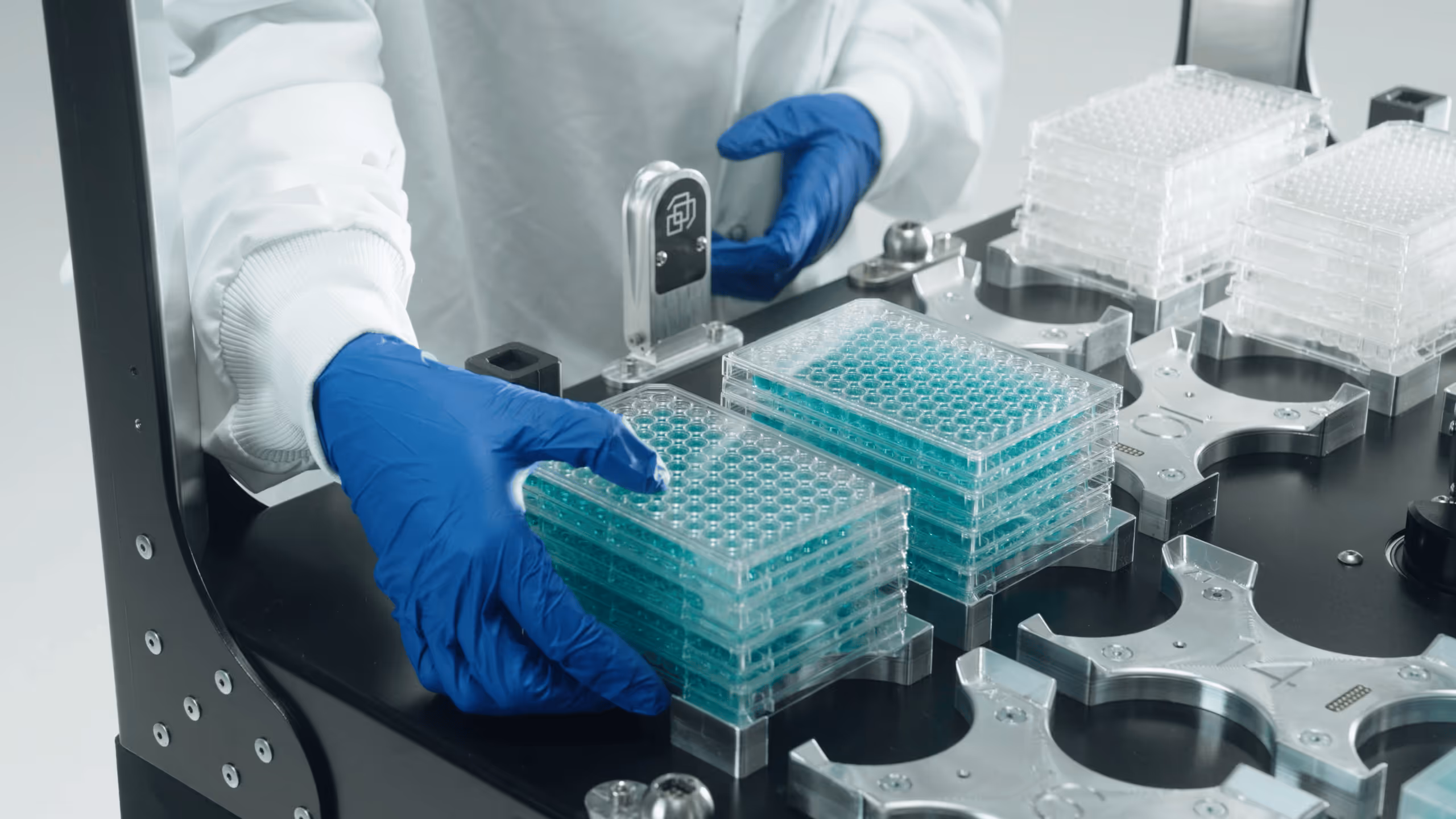 Gloved hands loading stacked multiwell plates onto an automated lab platform as part of an integrated biology lab system