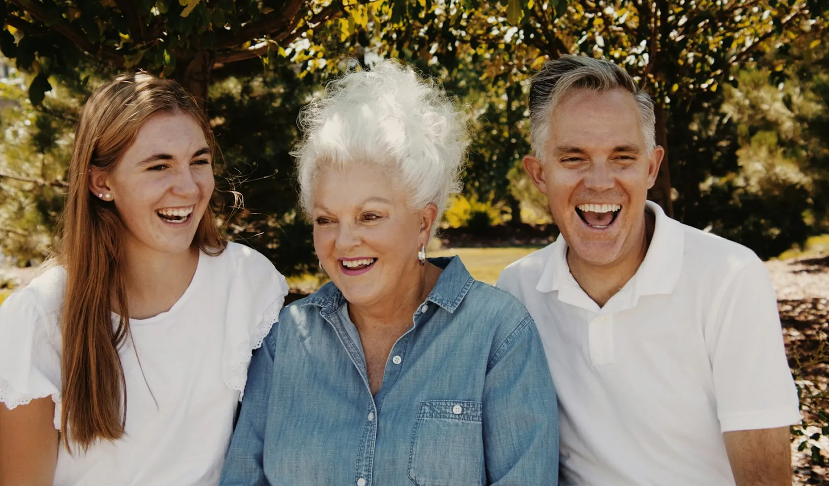 Three people, a young woman, an older woman, and a middle-aged man, smiling and sitting outdoors with trees in the background.